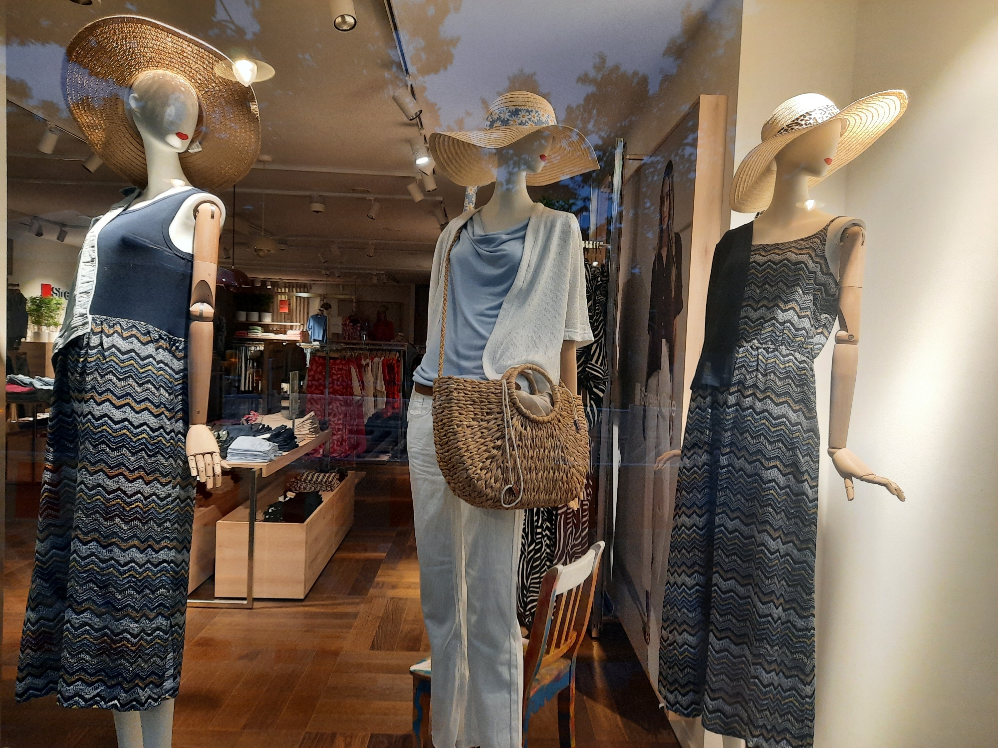 A storefront window displays three mannequins dressed in summer fashion. The mannequin on the left wears a dark blue sleeveless top over a long, patterned skirt and a wide-brimmed straw hat. The center mannequin is dressed in light blue pants, a light-colored cardigan, and a straw hat adorned with daisies. It holds a woven handbag. The mannequin on the right wears a patterned sleeveless dress and a straw hat with a leopard print band. The mannequins are posed in front of a store interior with clothing racks and display shelves. The floor is wooden, and the ceiling has track lighting. Reflections of trees and sky are visible on the window. The store appears to be located in Rosenheim, Germany.