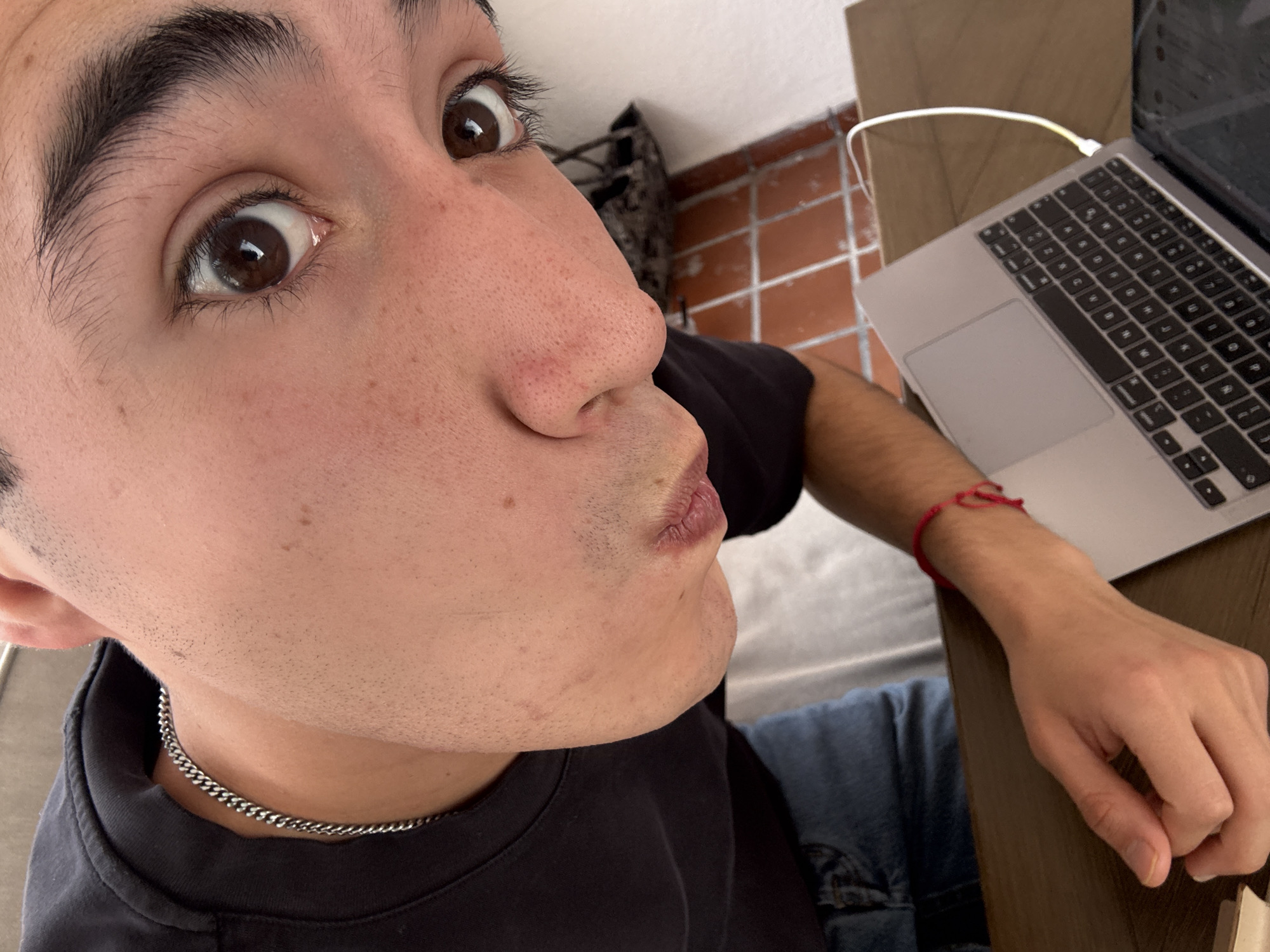 A man takes a close-up selfie, puckering his lips. His right eye is wide open, revealing a brown iris. He is wearing a black shirt and a silver chain necklace. His left hand rests on a wooden table next to an open laptop, which is plugged into a charger. A red string bracelet is visible on his wrist. In the background, there is a tan, brick-like wall. The location cannot be confirmed by the image, but the prompt mentioned Puebla, Mexico.