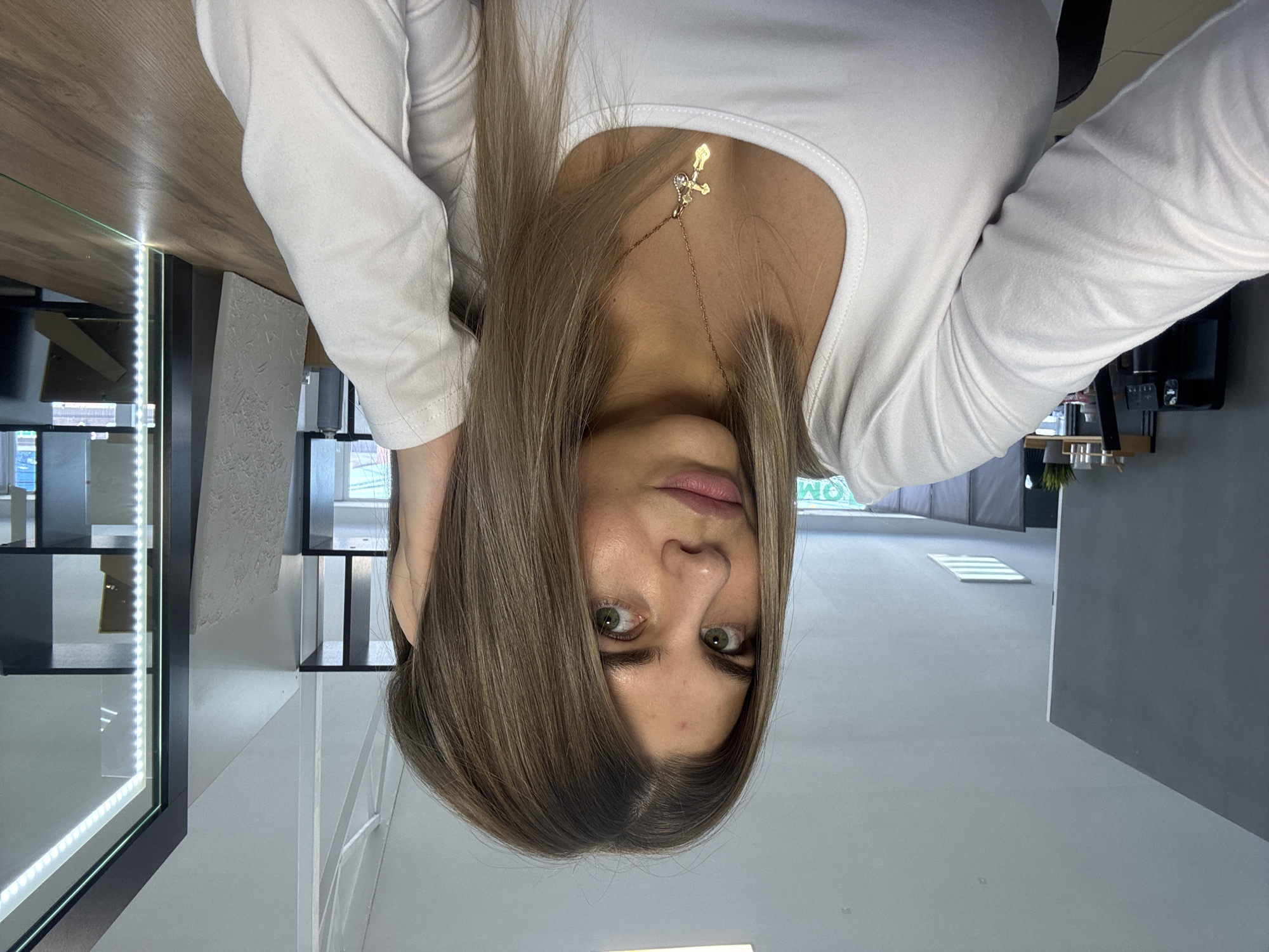A woman with shoulder-length blonde hair is looking down at the camera. She is wearing a white, long-sleeved shirt and a gold necklace with a cross pendant. The location is an indoor space in the city of Chaikovsky, Russia. Visible in the background are a table, a glass display case with LED lighting, a white wall, and a gray wall.