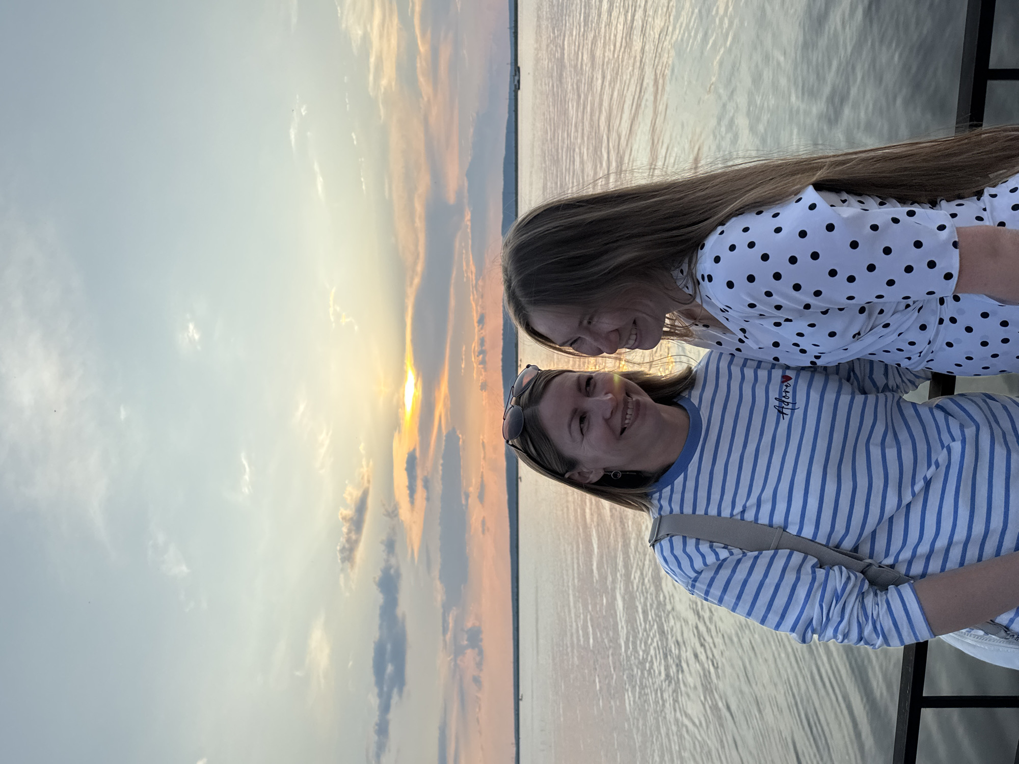 Two women are standing on a pier at sunset, looking at each other and smiling. The woman on the left is wearing a blue and white striped long-sleeved shirt and sunglasses on her head. The woman on the right is wearing a white dress with black polka dots. The sun is setting over a large body of water, with a hazy orange and yellow sky. The location is the embankment of the Kama River in Chaikovsky, Russia.