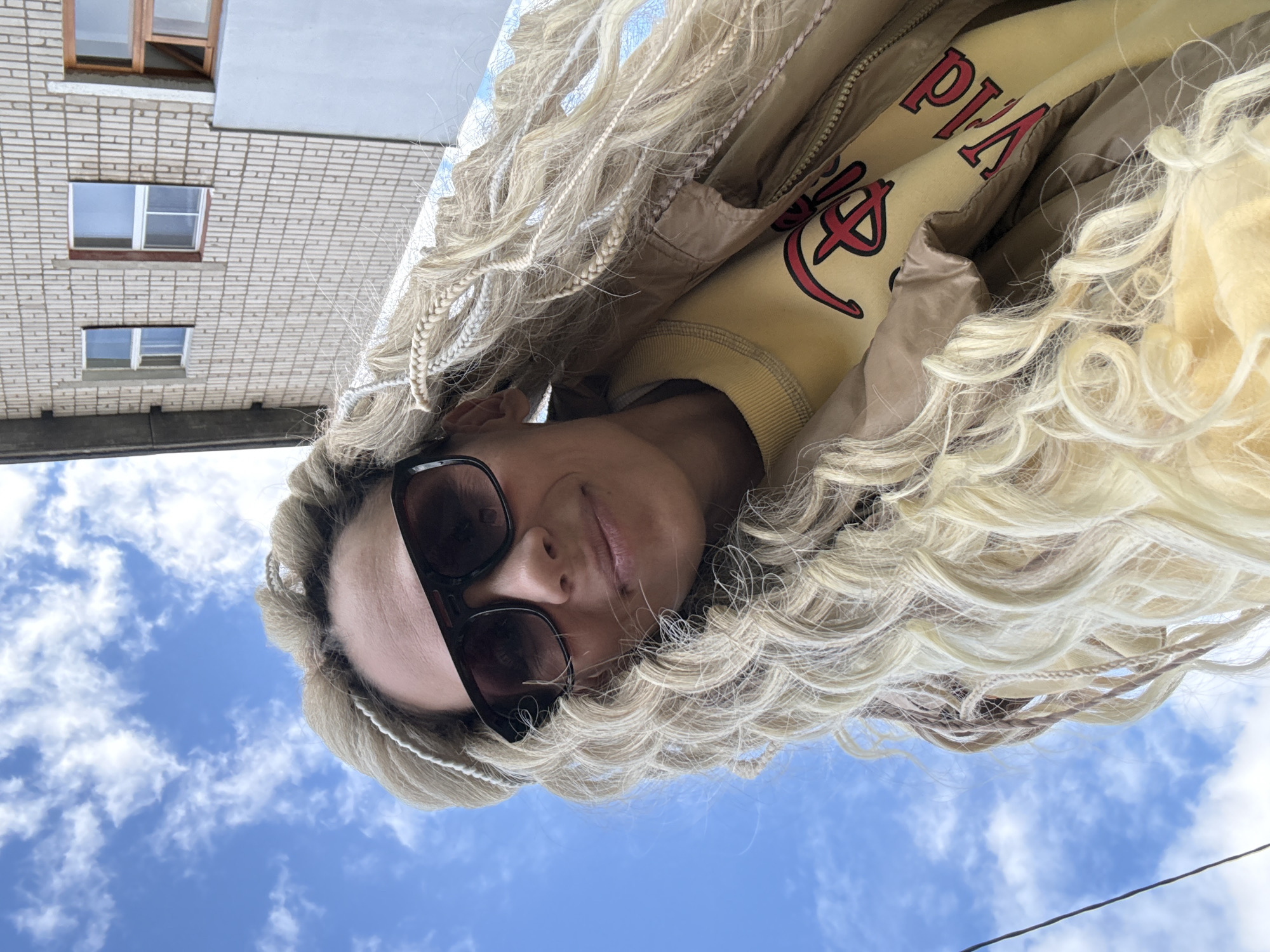 A woman with blond braided hair is seen from a low angle, wearing sunglasses and a yellow sweatshirt with a red design under a brown jacket. Behind her is a building with multiple windows and a clear blue sky with scattered white clouds. The location is Rusanovo, Russia.