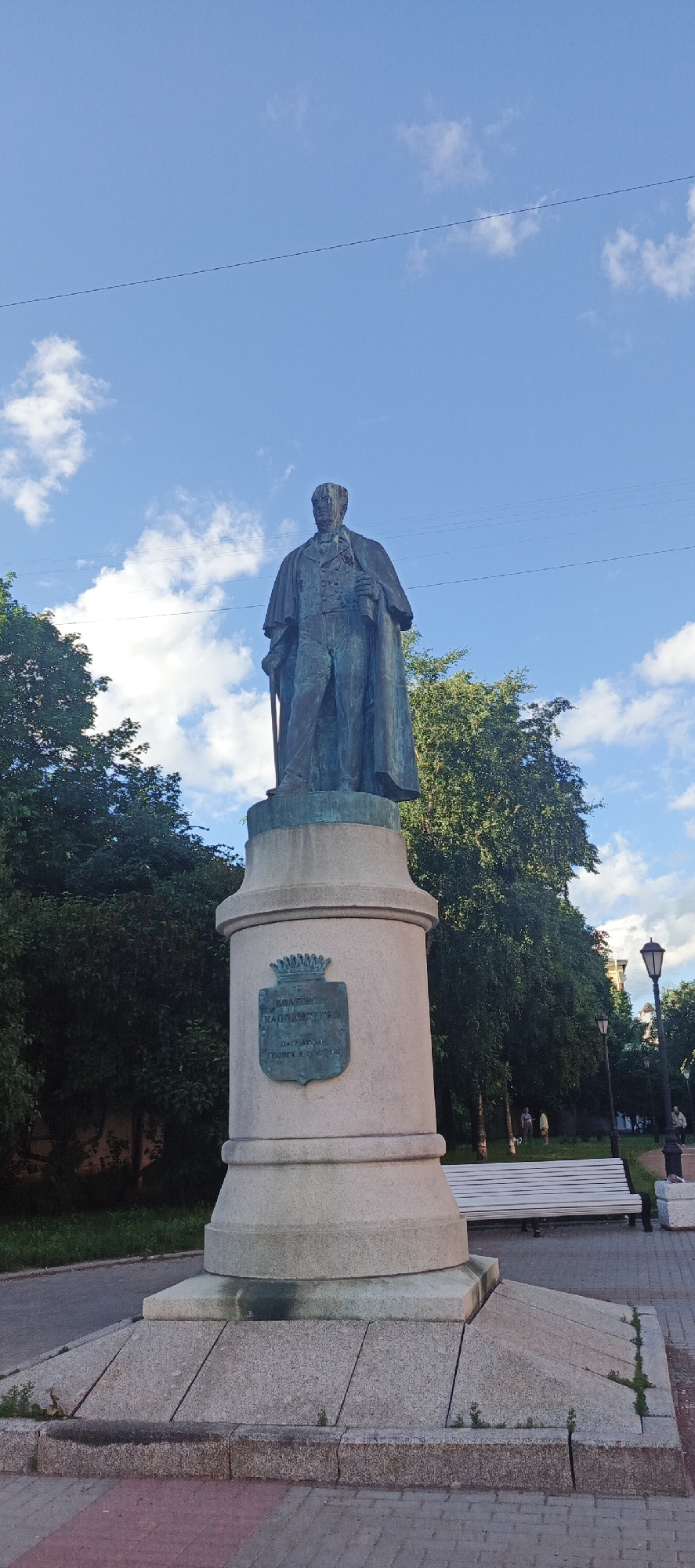 A bronze statue of a man in a long coat and suit stands on a stone pedestal in a park. The man holds a cane in his left hand and has his right hand in his pocket. A coat of arms with a crown is affixed to the front of the pedestal, bearing the inscription in Cyrillic: "КАДЕТСКОМУ КОРПУСУ 1857". Above this, there is another inscription, also in Cyrillic, which appears to be part of the same dedication.
The statue is set against a bright blue sky with scattered white clouds. Lush green trees frame the sides of the statue, and a paved walkway with red and grey bricks is visible in the foreground. In the background, a white park bench, lampposts, and a few people walking can be seen. The scene appears to be set during the daytime, possibly late afternoon or early evening, with the sun casting a gentle light. The overall atmosphere is serene and peaceful. The presence of a park bench and people suggests it is a public space designed for leisure and remembrance. The statue's location context is Saint Petersburg, Russia.