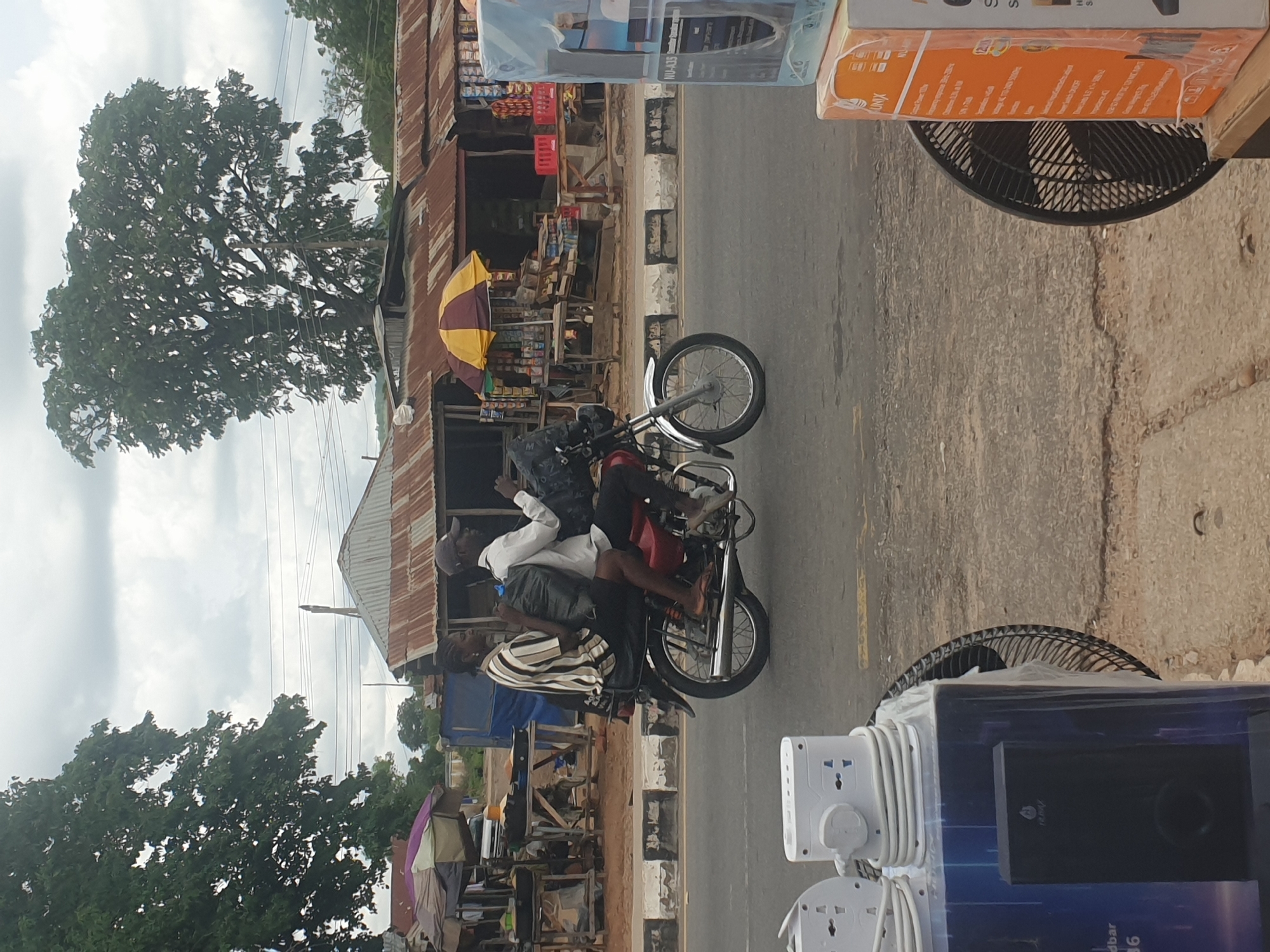 The image shows a street scene in an urban area of an Unknown city. Three individuals are riding on a motorcycle on the right side of the road. Adjacent to the road are shops and stalls, covered with awnings and umbrellas. Various objects, like a fan and electronics, are partially visible in the foreground. Large trees are visible in the background under an overcast sky.