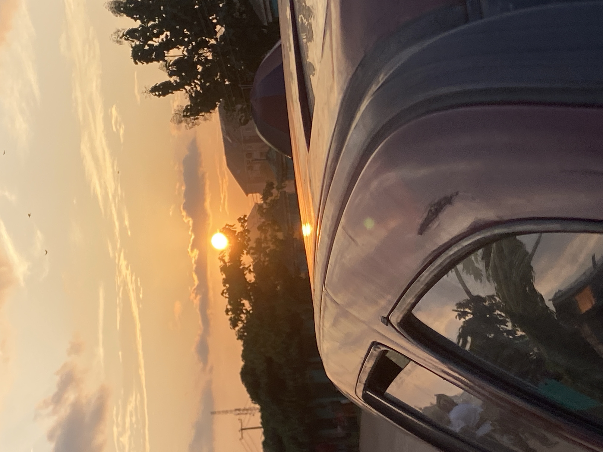 A sunset with the sun partially obscured by clouds is visible in the sky over Abakaliki, Nigeria. The sun casts an orange glow across the sky, illuminating scattered clouds. In the foreground, the roof and side of a dark-colored vehicle are visible, reflecting the sunset. Trees and buildings are silhouetted against the sky in the background. A utility pole with wires is also present in the distance.