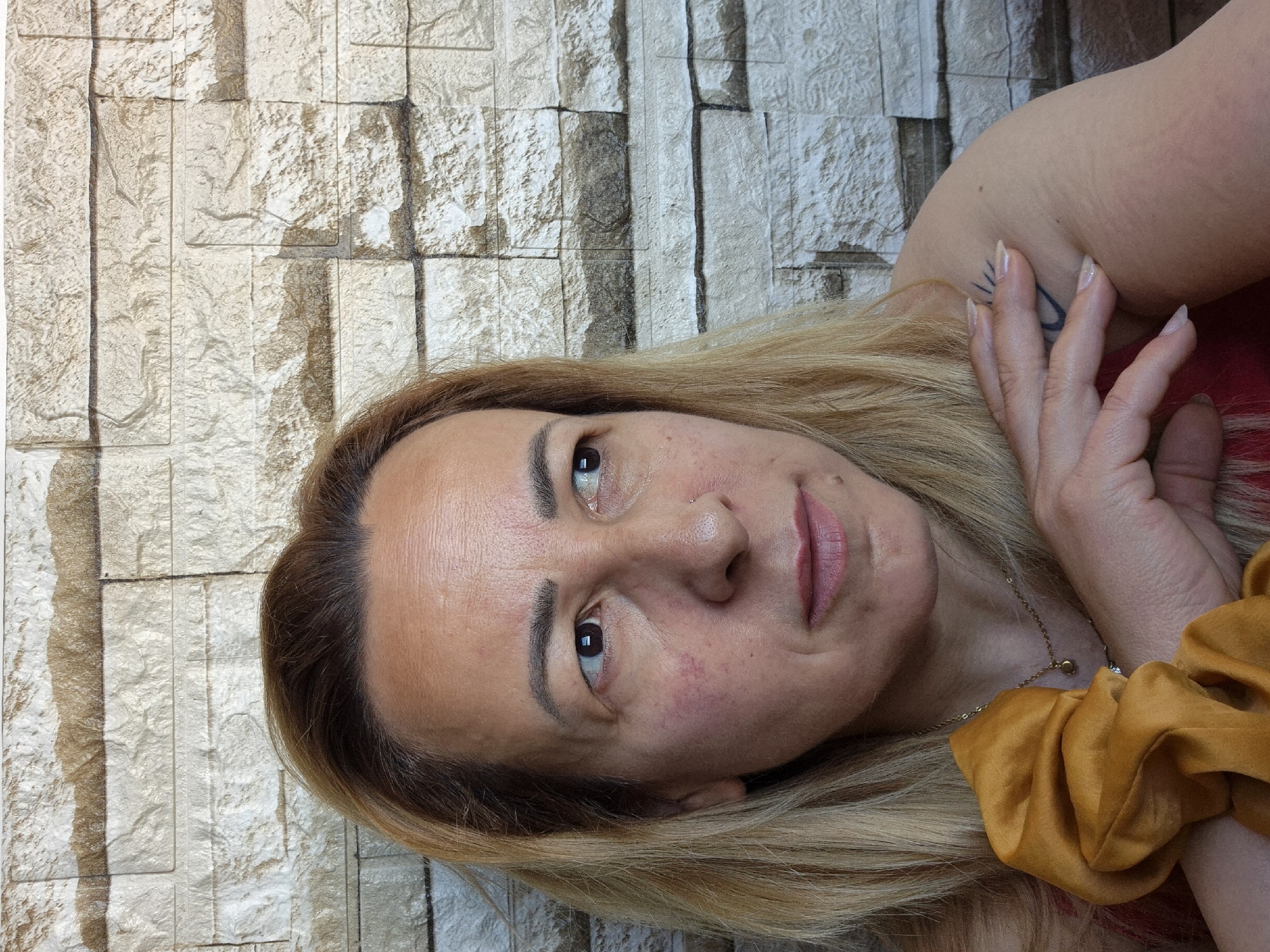 A medium close-up, portrait-oriented photograph features a woman with blonde hair and a fair to medium skin tone, positioned against a textured stone wall. Her head is tilted, giving the impression that she is lying down, and she is looking directly towards the viewer.
The woman's hair is long and blonde, with darker roots visible at the hairline. Her facial features include arched eyebrows, brown eyes, and a neutral expression with a slightly closed mouth. Her skin shows some redness, particularly on her cheeks. She has a small nose piercing on her right nostril.
She is wearing a red garment, partially visible on her shoulder and arm. Her left arm is bent, with her hand resting near her head. Her fingernails are long and manicured with a light polish. On her left shoulder/upper arm, a dark tattoo, possibly depicting a stylized sun or a partial symbol, is visible. Around her neck, a thin gold chain necklace with a small pendant is discernible. A large, mustard yellow, satin or velvet scrunchie is worn on her wrist or held in her hand, resting on her hair.
The background consists of a light-colored stone wall, composed of irregular, rectangular blocks with prominent texture and varying shades of beige, gray, and brown. The lighting appears bright and natural, casting subtle shadows that highlight the textures of the wall and her features. There is no visible text.