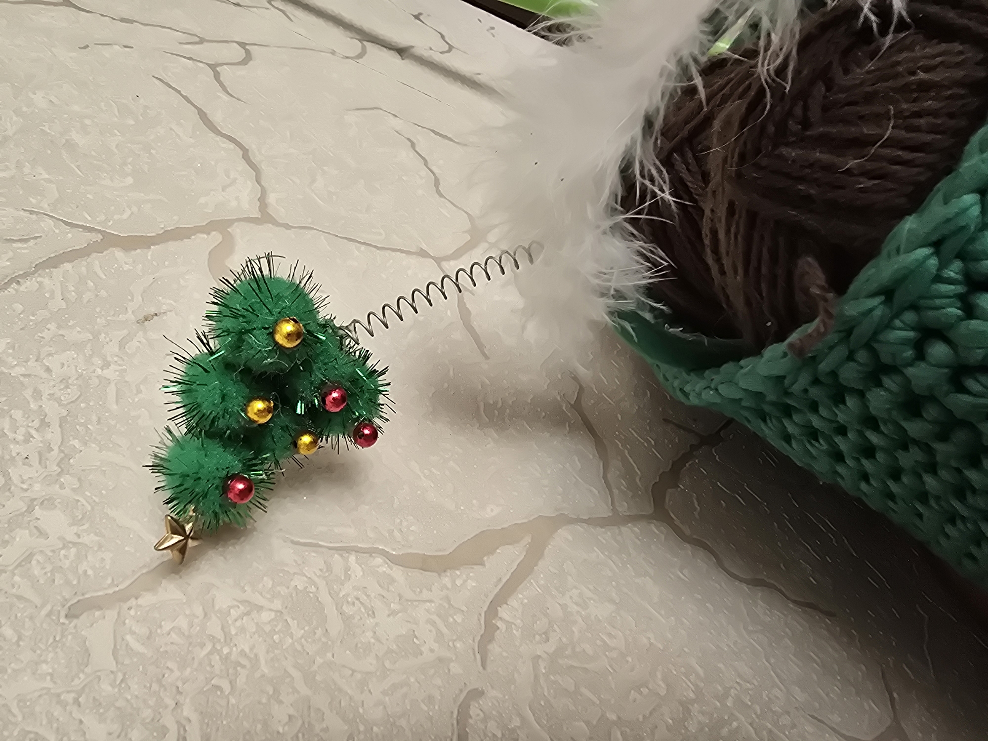 The image shows a close-up of a small, artificial Christmas tree topper, adorned with red and gold baubles and a gold star. It is attached to a coiled spring and has a fluffy white trim at its base. The topper appears to be part of a larger decorative item, possibly a Christmas stocking, which is partially visible at the bottom of the frame, filled with brown yarn. The background is a textured, light-colored wall with a faint pattern resembling tree branches or cracks. The scene is domestic, with no identifiable landmarks or context suggesting a specific location in Krasnoyarsk, Russia.
