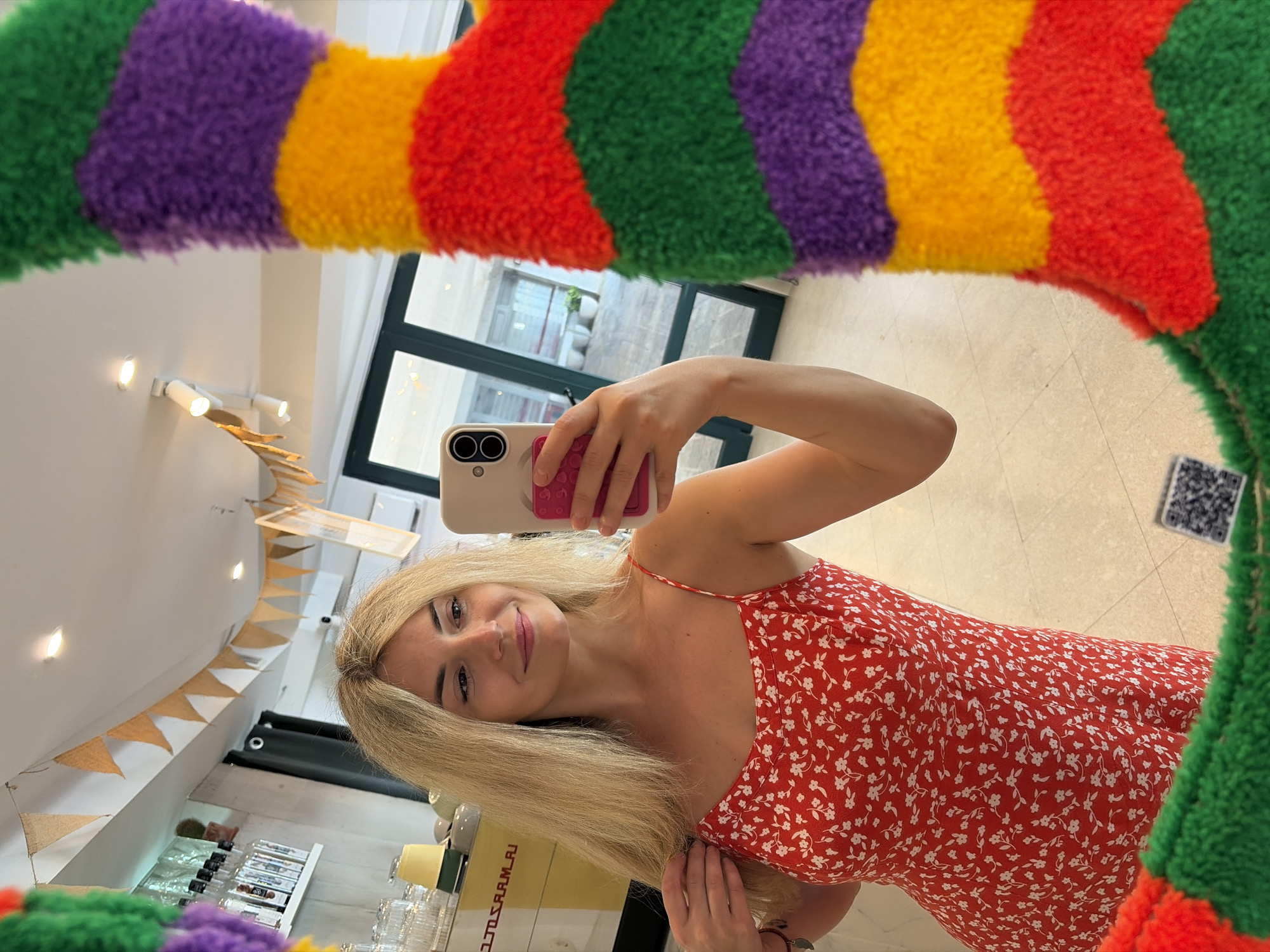 In Budva, Montenegro, a young blonde woman with fair skin and light makeup takes a mirror selfie. She is wearing a red dress with a white floral pattern and spaghetti straps. Her hair is long and slightly tousled. In her right hand, she holds a white smartphone with a pink pop socket on the back. The background suggests an indoor establishment, possibly a cafe or shop, with shelves stocked with bottles and glassware visible. Bunting, in a light brown, triangular shape, is strung across the ceiling. A brightly colored, fuzzy mirror frame, featuring stripes of purple, yellow, red, and green, partially obscures the right side of the image. A QR code is visible on the floor in the lower right corner. The lighting appears to be artificial, with spotlights on the ceiling.