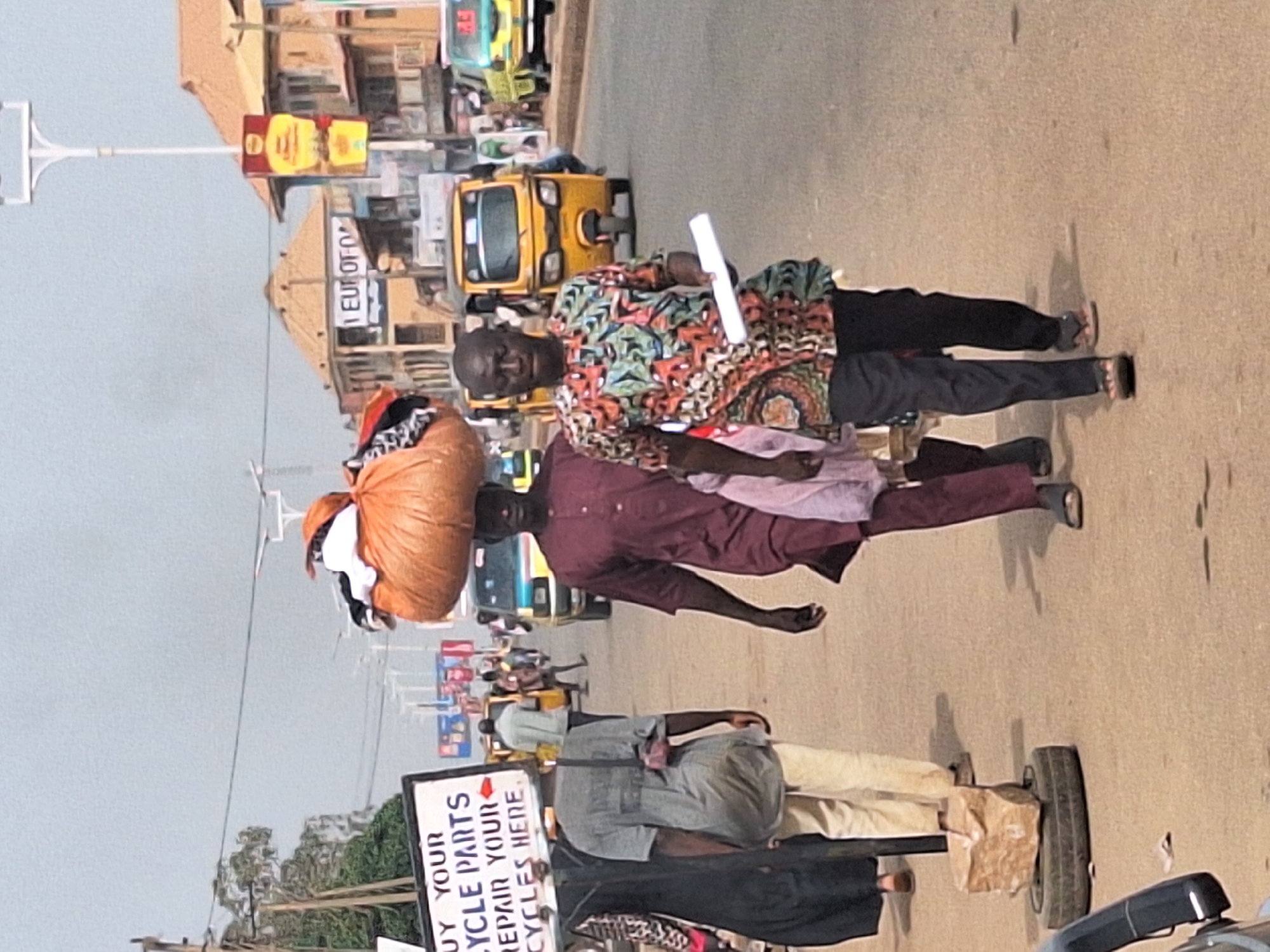 In Sabon Tasha, Nigeria, the image captures a street scene with pedestrians and vehicles. A man balances a large sack of clothes on his head. Another man, beside him, holds a rolled-up white paper. A tricycle is visible behind them. Further down the street, a person pulls a cart with a sign advertising bicycle parts and repair. Various other pedestrians and vehicles populate the scene. Buildings and commercial signage are visible in the background.