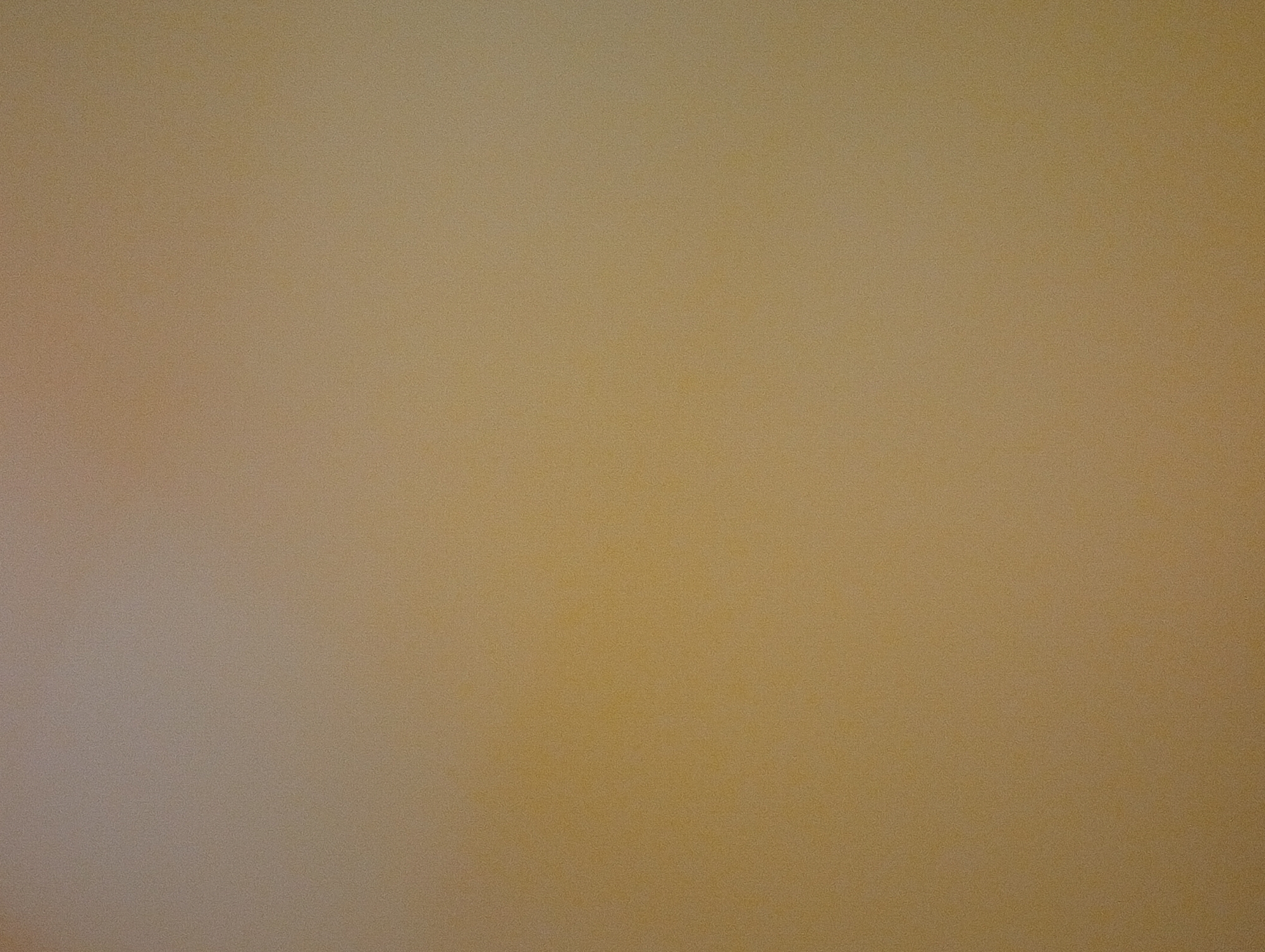 The image is a photograph of a wall with a textured surface. The color of the wall is a light brown or beige. The lighting in the image creates a gradient, with the left side appearing lighter and brighter, possibly due to a light source off-camera, and the right side being slightly darker. The surface of the wall has a subtle, uniform grain or texture visible throughout. There are no discernible objects, people, or actions depicted in the image.