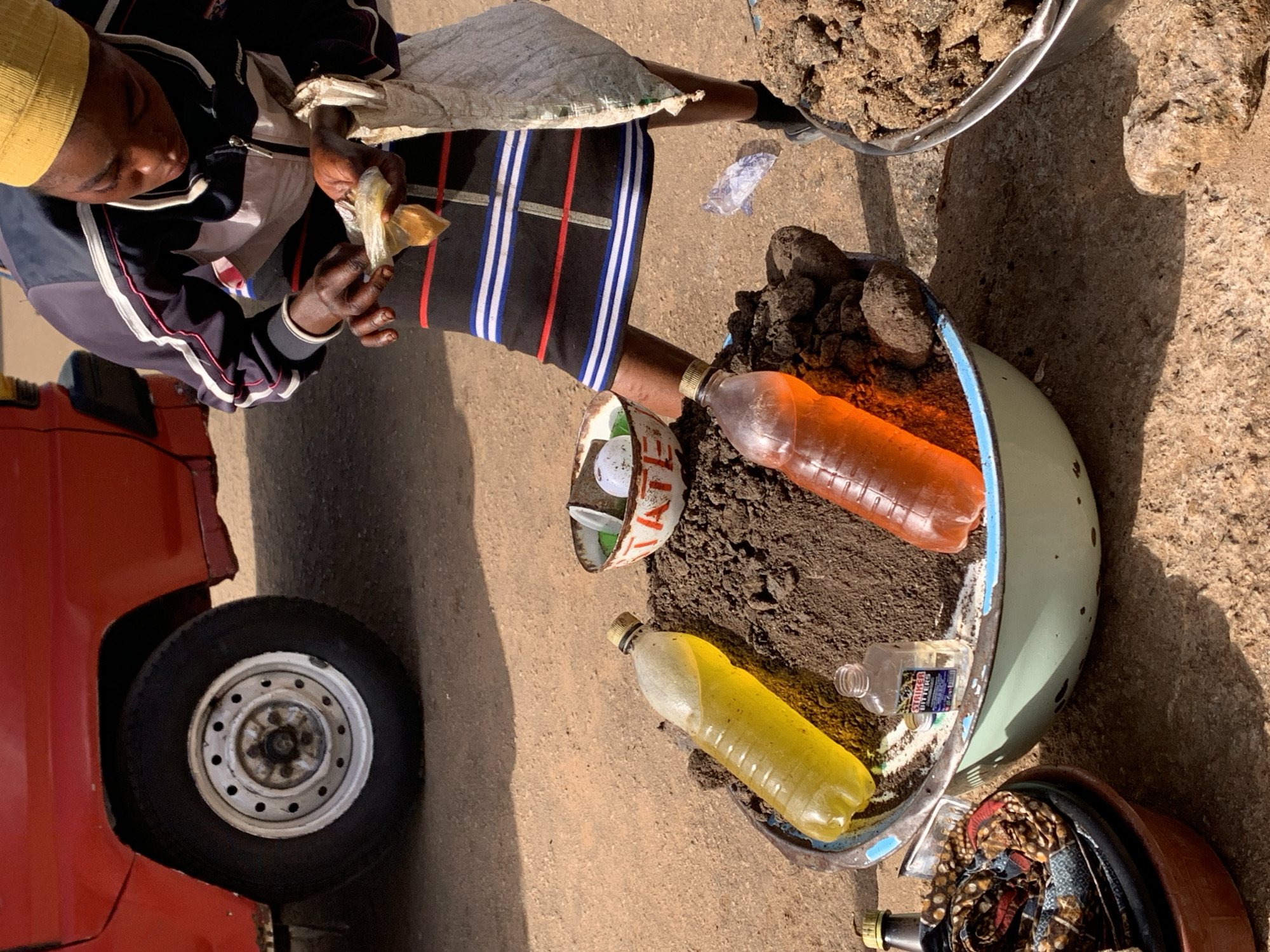 A person wearing a yellow head covering and a jacket with white and pink stripes is hunched over, holding a small, light-colored object in their hands. They are wearing striped pants, with blue, white, and red stripes. The scene appears to be outdoors, with a red vehicle visible to the left, including a tire. In the foreground and to the right, there are several large, shallow bowls or containers. One light blue-green bowl contains dark soil and two plastic bottles, one filled with orange liquid and another with yellow liquid. Another, darker bowl below it contains fabric with a patterned design. A large, rough stone is visible to the right of the bowls. The ground is a mix of dirt and concrete. The lighting suggests it is daytime. The overall impression is that of a market stall or a roadside vendor.