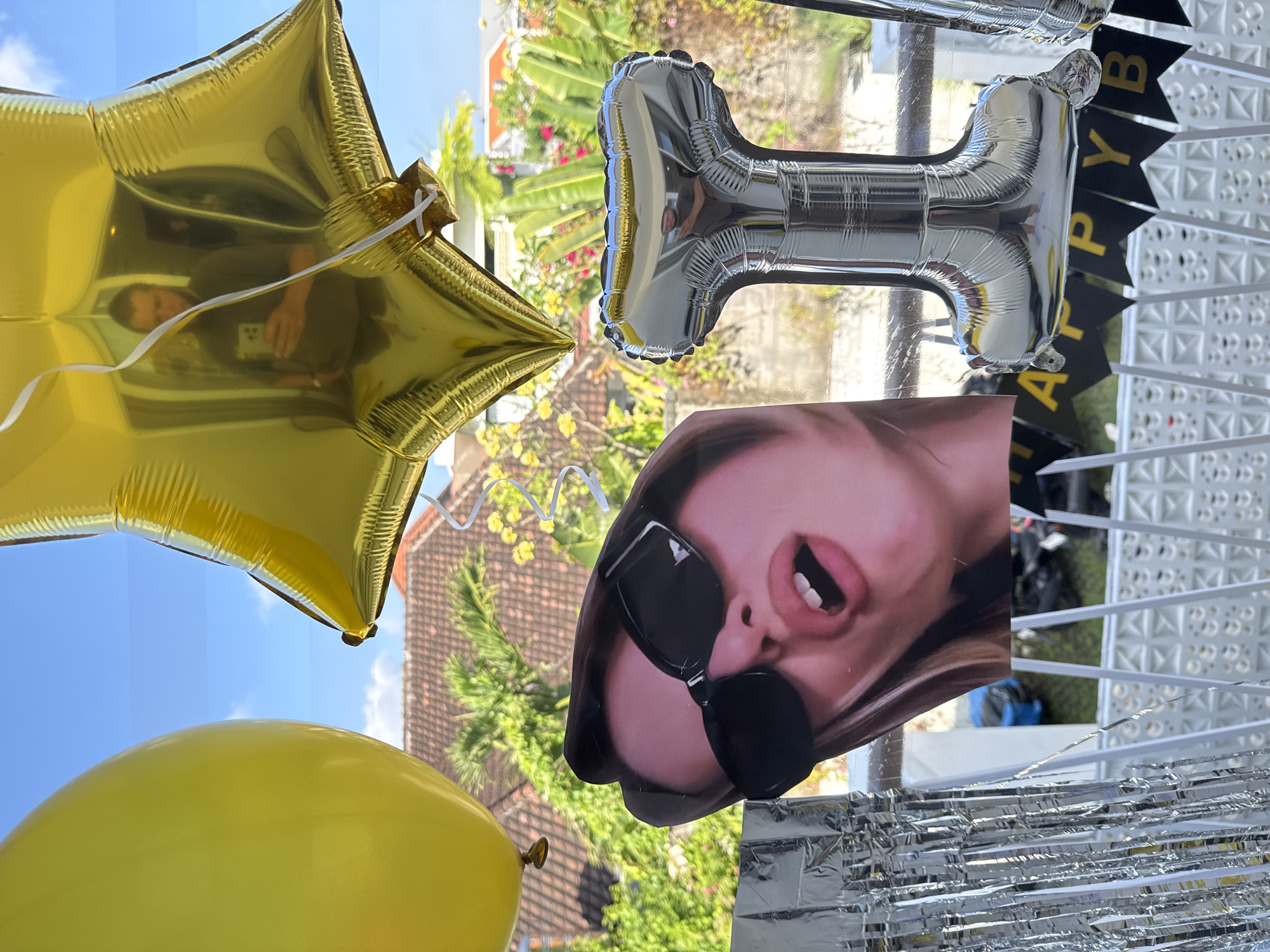 A close-up shot features several balloons and a cutout of a woman's face, set against a backdrop of a tropical outdoor scene visible through a window. In the foreground, a large yellow balloon occupies the left side of the frame, partially obscuring a gold star-shaped balloon positioned above and to the right. A cutout of a woman's face, wearing oversized black sunglasses and with her mouth open, is placed in the center-left. To the right of the cutout is a silver foil balloon shaped like the number "1". Below the balloons and cutout, a black banner with yellow lettering spells out "HAPPY BIRT". The background shows lush green foliage, a terracotta-tiled roof, and a bright blue sky with scattered clouds, indicative of a sunny day in a tropical location. The scene appears to be part of a celebration, possibly a birthday, taking place in a residential or hospitality setting in North Kuta, Indonesia.