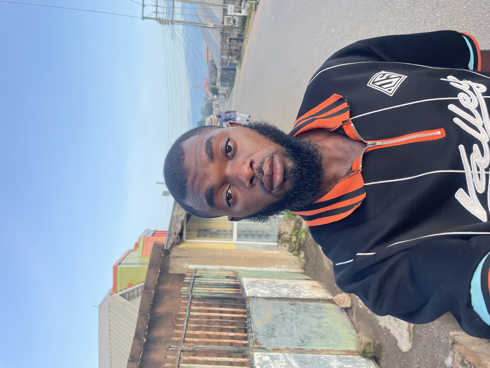 In Jos, Nigeria, a dark-skinned man with a beard and short hair stands in the foreground, facing the camera. He is wearing a black polo shirt with orange and white stripes on the collar and down the front, featuring a zipper closure. The background shows a street scene with buildings on either side, power lines overhead, and a few people walking in the distance. To the left, a rusty metal gate and a wall are visible. The sky is clear and blue.