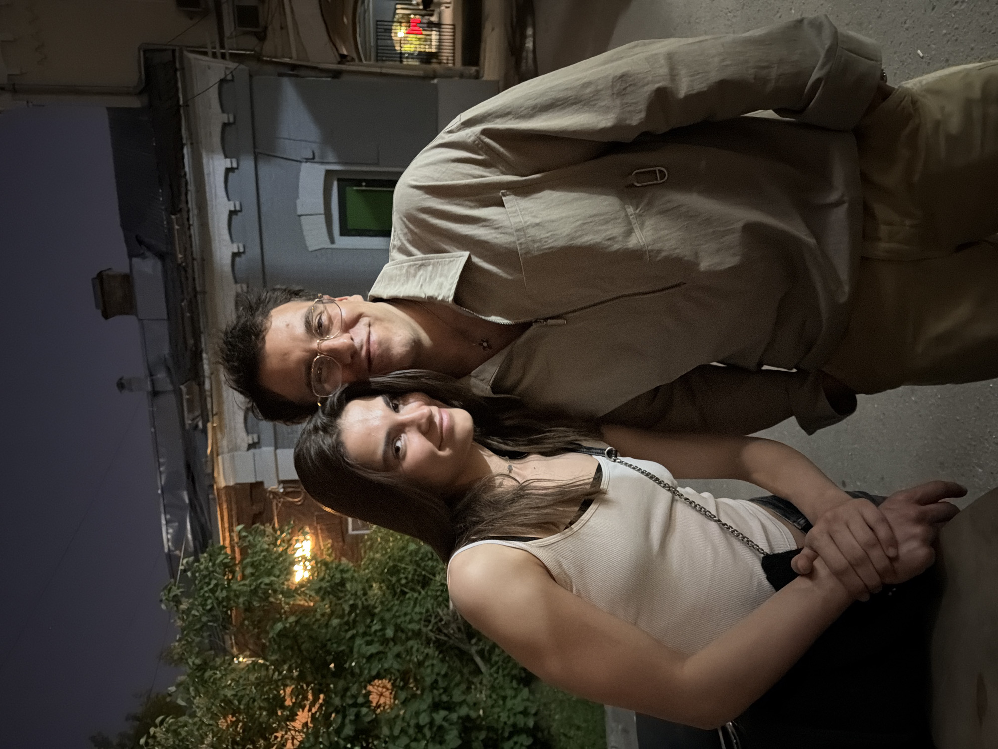 A medium shot shows a young woman and a man standing close together at night, outdoors. The woman, on the left, has long dark hair and is wearing a light-colored tank top and a black bra. She is holding the man's hands, which are resting on her upper arm. A black purse with a chain strap is visible over her shoulder. The man, on the right, has short dark hair and is wearing glasses, a light-colored collared shirt with a zippered front, and khaki pants. He has his left hand in his pants pocket and his right arm is around the woman's shoulders, but not fully visible.
Behind them, a building with a dark roof and light-colored walls is visible. A large green tree with illuminated foliage is to the left of the couple. The background appears to be a street scene in Moscow, Russia, possibly near a residential or commercial area with old architecture. The lighting suggests it is nighttime, with artificial lights illuminating the tree and a muted glow from the sky.