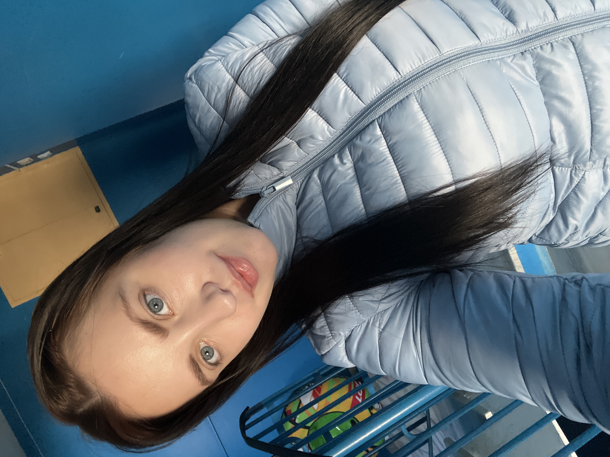 A woman with long dark hair and light blue eyes is looking at the camera. She is wearing a light blue quilted jacket. The background is a blue wall and floor with a yellow box in the corner. There is a blue metal railing with a colorful ball visible behind it. The location is presumably Марковский, Russia.
