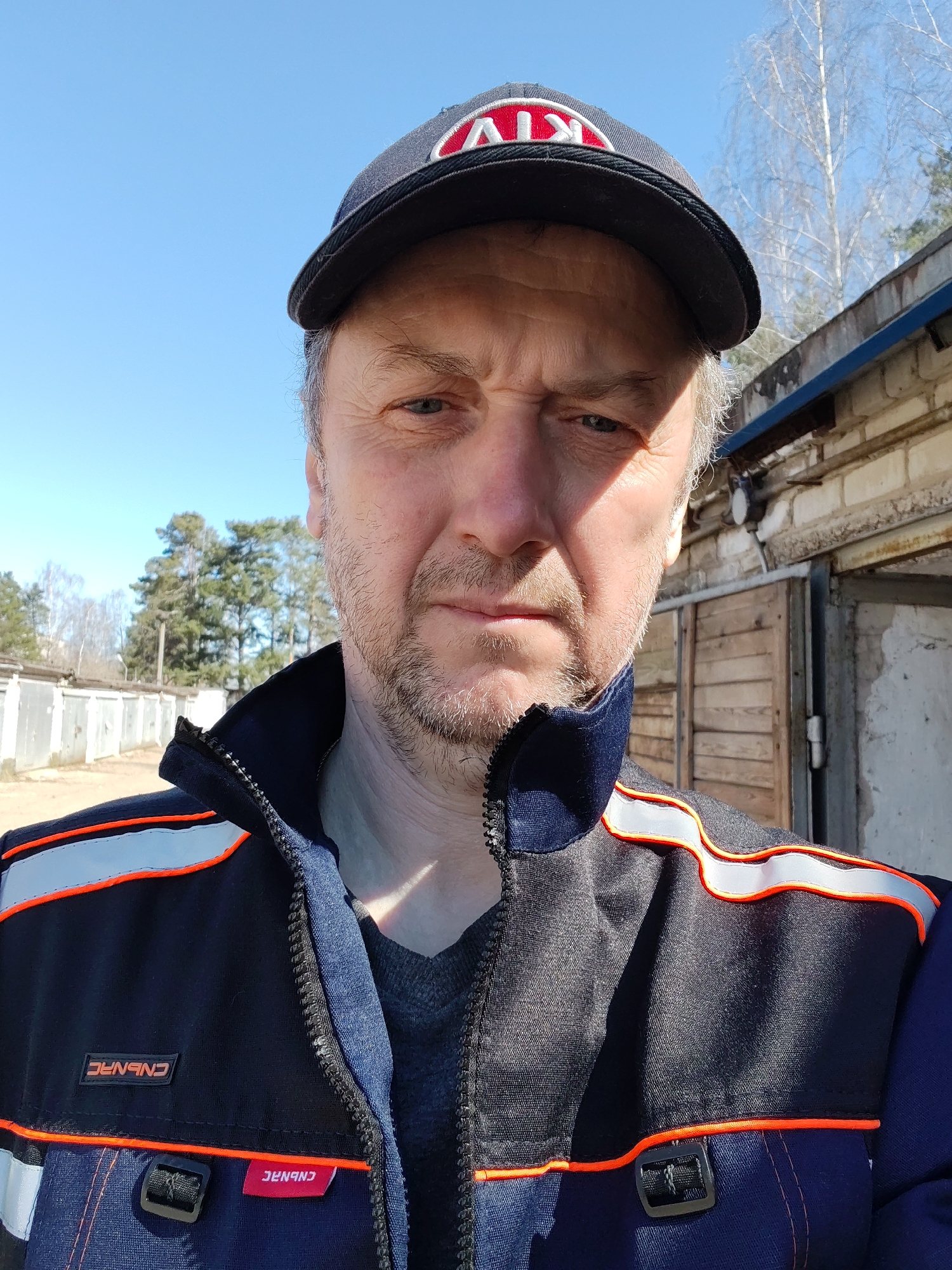 A middle-aged man with a light beard and wearing a dark baseball cap with the letters "AVK" in red and white, and a dark blue jacket with orange and white reflective stripes, poses for a selfie. The jacket has a small rectangular patch with white text in a red box that appears to be a brand label. Behind him is a concrete structure, possibly a garage, with a wooden door. In the background, trees with bare branches and a clear blue sky are visible. The man is the primary subject and is looking directly at the camera. The photograph was taken in Chaikovsky, Russia.