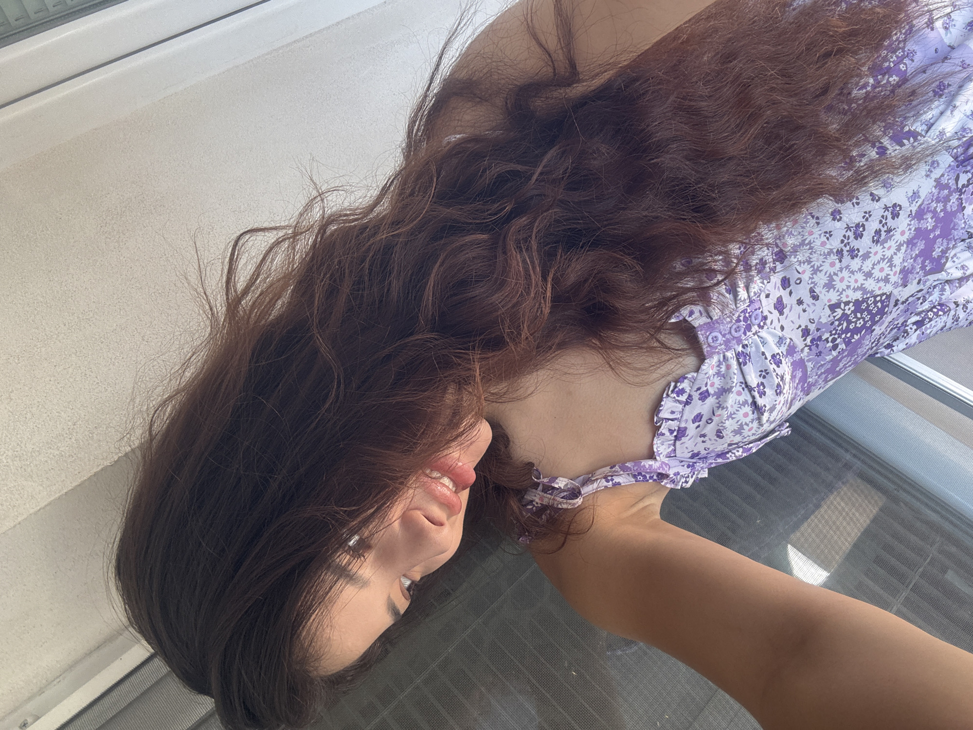 A low-angle, close-up shot depicts a person lying down, seemingly inside, with their head towards the lower left corner of the frame. The individual has long, wavy brown hair that spreads out across the top of the image. They are wearing a light purple floral print dress with ruffled trim. Only a portion of their arm and upper body are visible. The background consists of a white wall and a window with a screen. The overall setting appears to be an indoor space. It is not possible to determine the specific location in Plovdiv, Bulgaria from the given information.