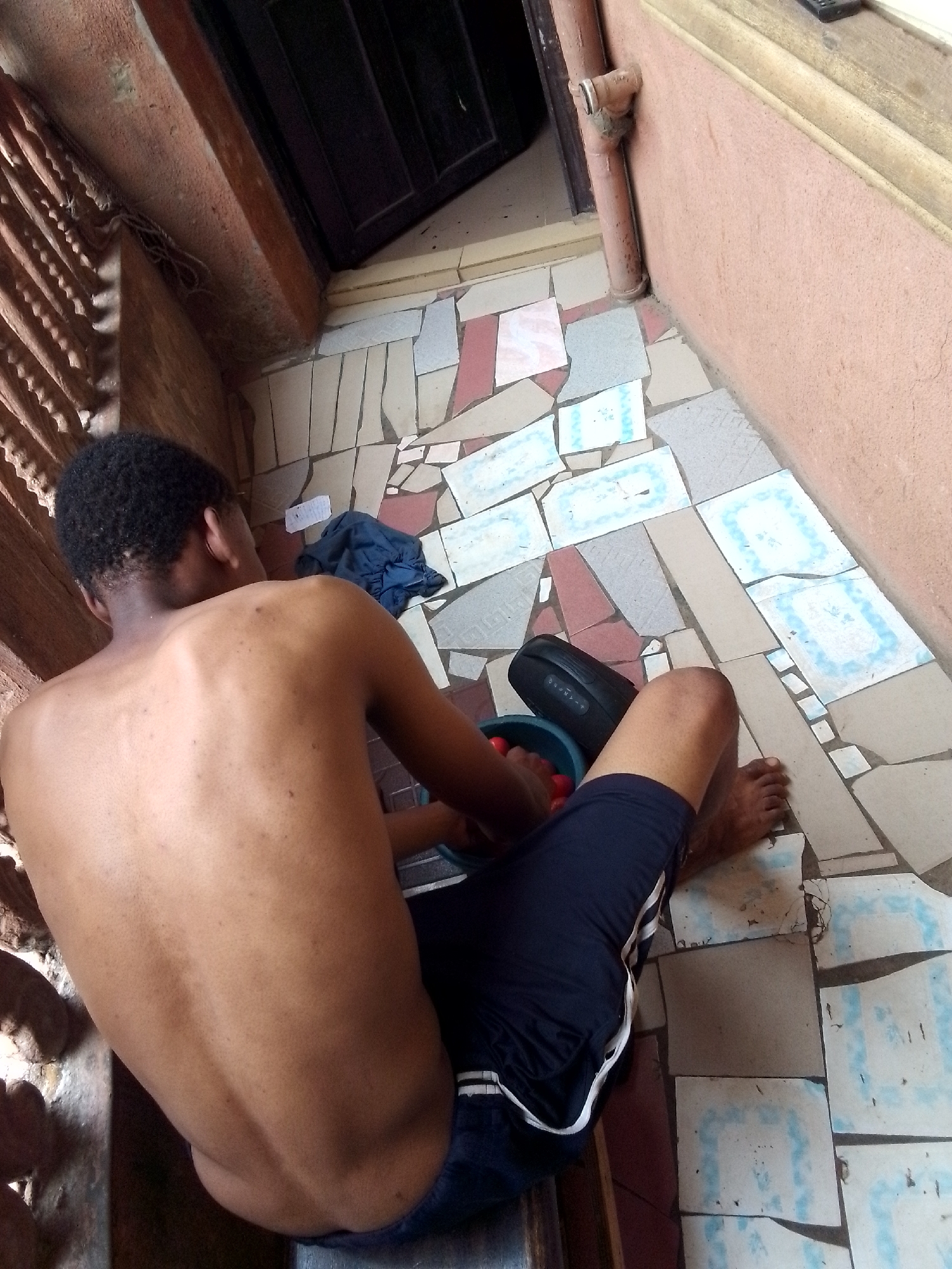 A high-angle shot shows a partially bare-skinned person sitting on a ledge or step in what appears to be an outdoor or semi-outdoor space. The individual is facing away from the camera, exposing their back and arms. They are wearing dark shorts with a white stripe along the side. Next to them is a blue container with red spherical objects visible inside. A black object, possibly a speaker, is situated beside the container. The flooring is composed of mismatched tiles of varying colors and shapes, suggesting a mosaic or patchwork design. A closed door and a section of wall are visible in the background. The location is labeled as Unknown, Unknown.