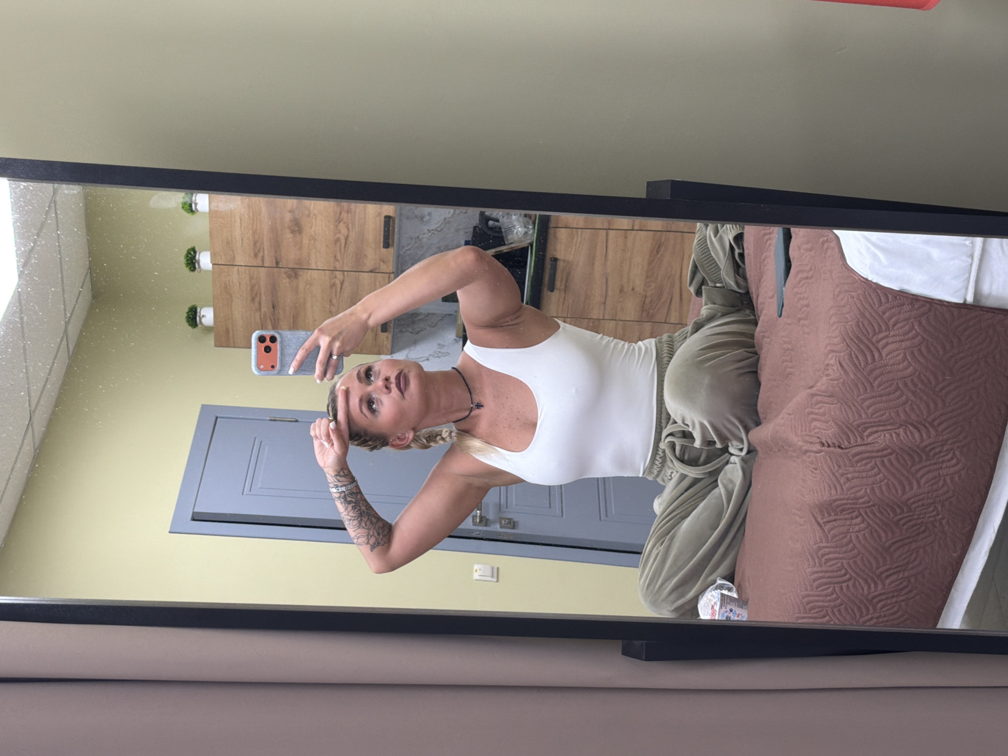 In a mirror selfie taken in Sarapul, Russia, a fair-skinned woman with blonde hair styled in braids is captured in an indoor setting. She wears a white tank top and light green sweatpants, and is adorned with a black necklace featuring a small cross-like pendant, a silver ring on her left hand, and multiple bracelets on her right wrist, which also displays intricate tattoos on her forearm. She holds a light grey smartphone with an orange side button in her left hand, taking the photo in a large, black-framed mirror. Her right hand is raised, touching her forehead, and she looks directly into the phone/mirror with a focused expression.
Reflected in the mirror behind her are elements of a room, likely a bedroom or living area, featuring light yellow walls. To her right (viewer's left in the reflection), wooden cabinets or drawers with dark handles are visible, topped with small potted plants. Further back, a light gray door with a prominent rectangular frame is seen, along with a light switch below it. To her left (viewer's right in the reflection), a bed is partially visible with a dark pink or brown quilted comforter and white sheets. A small, clear decorative object sits on a surface near the bed. There is no visible text in the image. The time of day and weather are not discernible from the indoor setting.