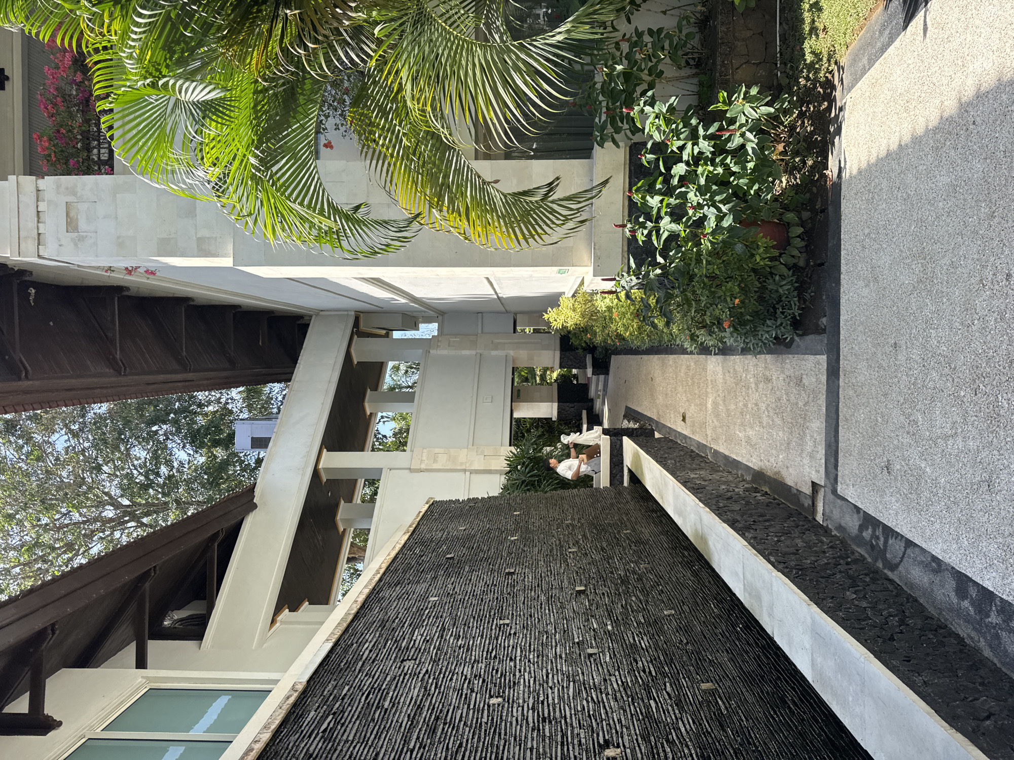 The image shows a man in a white shirt and brown shorts standing next to a dark stone waterfall wall, holding a white object. He appears to be performing some task, possibly cleaning or maintaining the area. The setting is an outdoor walkway with a pathway made of light-colored stone. To the right, there is a building with white stone facade and lush green palm trees and other plants. The architecture suggests a tropical resort or hotel in Kuta Selatan, Indonesia, characterized by open walkways, natural materials, and abundant greenery. The sun is shining, casting shadows on the pathway. In the background, there are more buildings with wooden eaves and balconies adorned with pink flowers.