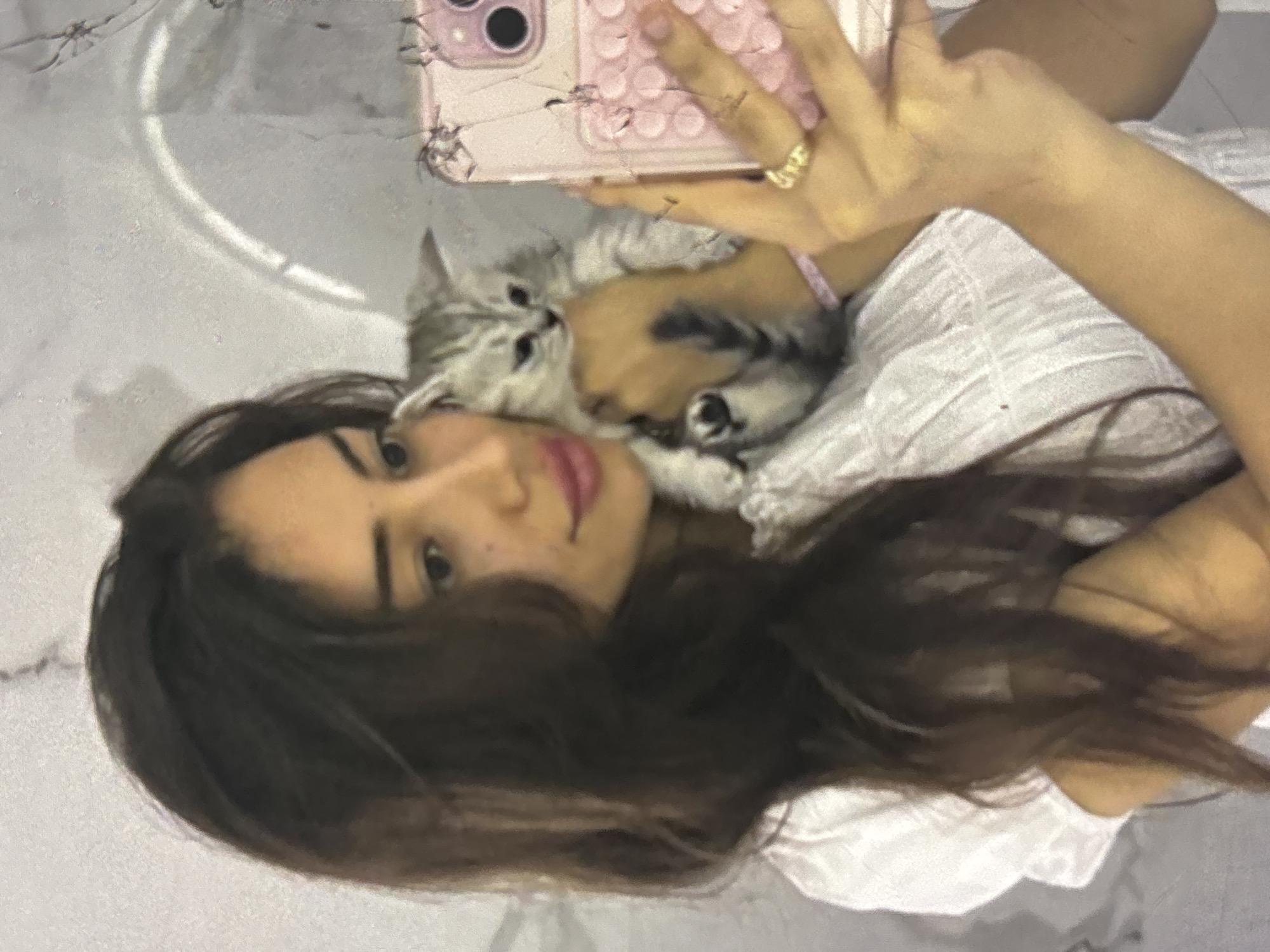 A young woman with long, dark hair, wearing a white dress, holds a small, grey and white striped kitten in her left arm while taking a selfie with her pink phone in her right hand. She is looking at her phone. A ring is visible on her right ring finger. The background appears to be a tiled wall, possibly in a bathroom or kitchen, with a light fixture reflected on the surface. The scene is set indoors, with no indication of the city of Orihuela, Spain.