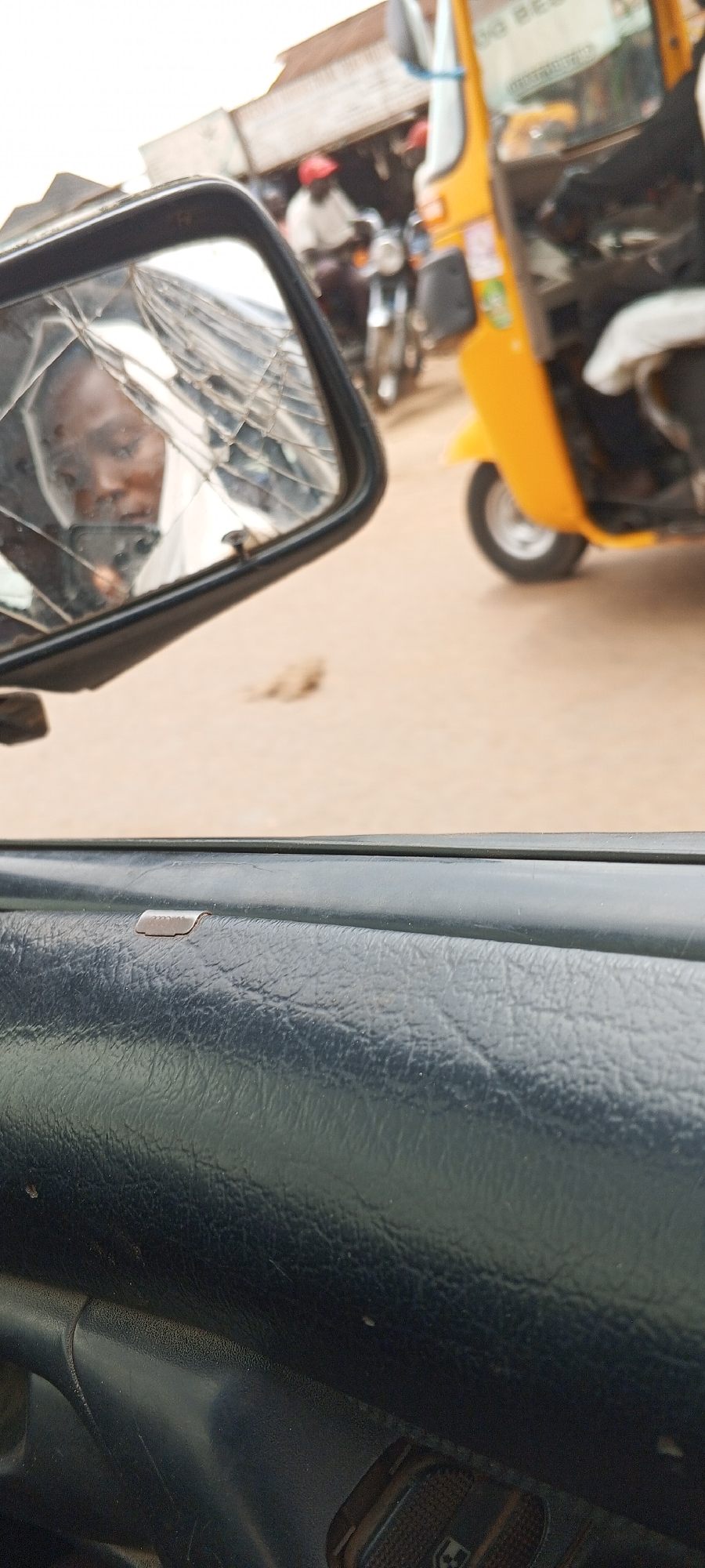 From within a vehicle in Masaka, Nigeria, the image shows a cracked side mirror reflecting a person with dark skin. In the background, partially obscured by the mirror and the vehicle's interior, are a yellow auto-rickshaw and a motorcycle with a rider wearing a red helmet. The environment appears to be an urban street scene. The interior of the vehicle is visible, showing part of the door panel.