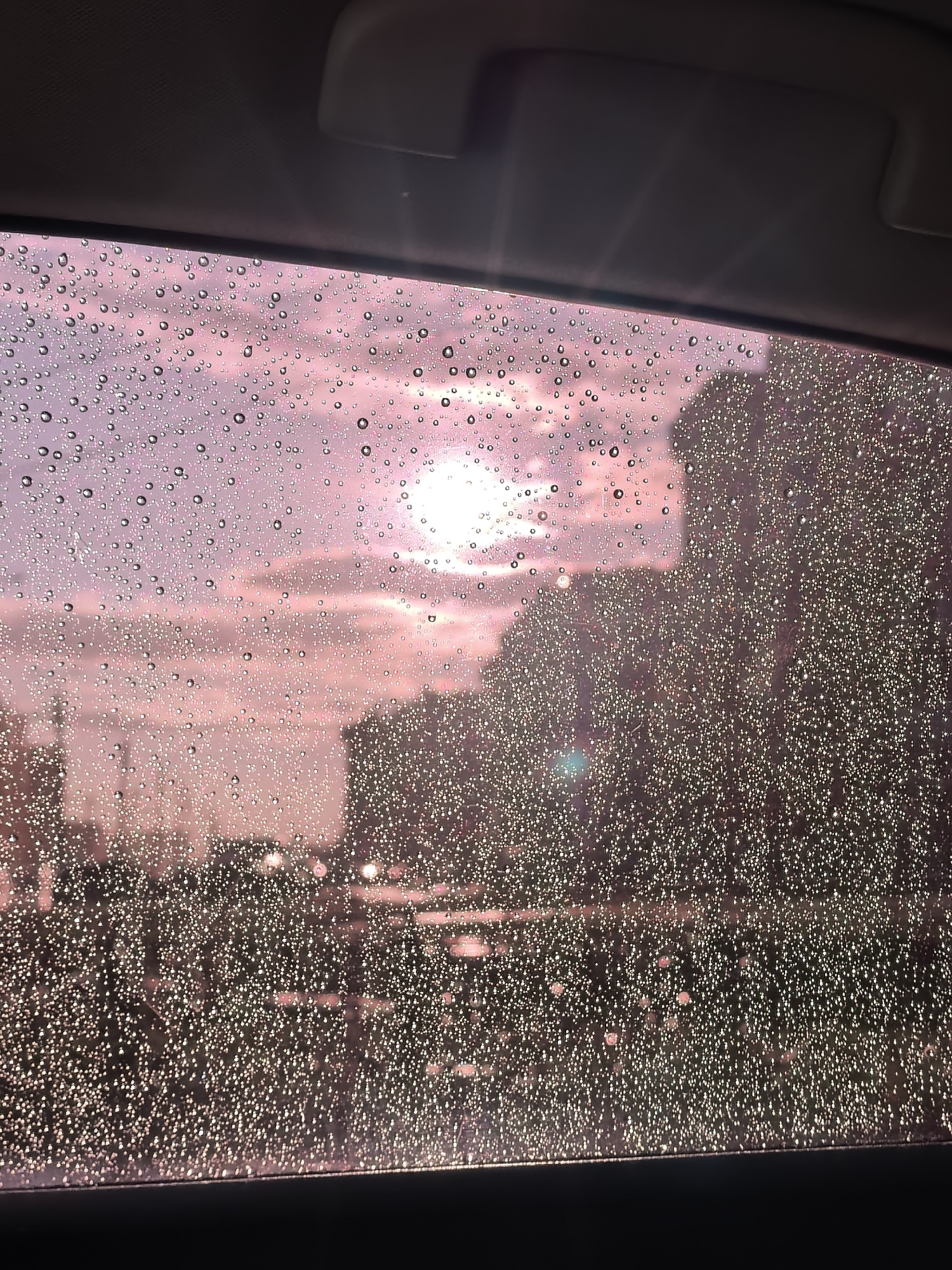The image shows a view through a window covered in raindrops, looking out at a cloudy sky with the sun partially visible. The sky is a pale pinkish-purple color. In the distance, silhouetted trees and buildings can be seen, appearing dark against the sky. The foreground is dominated by the water droplets on the windowpane, which distort the view and reflect light. The interior of a vehicle is visible at the top of the frame, with a dark gray headliner and a sun visor. There are no people or specific landmarks visible to indicate the location in Sterlitamak, Russia. The scene appears to be a transient moment captured during or after rainfall.