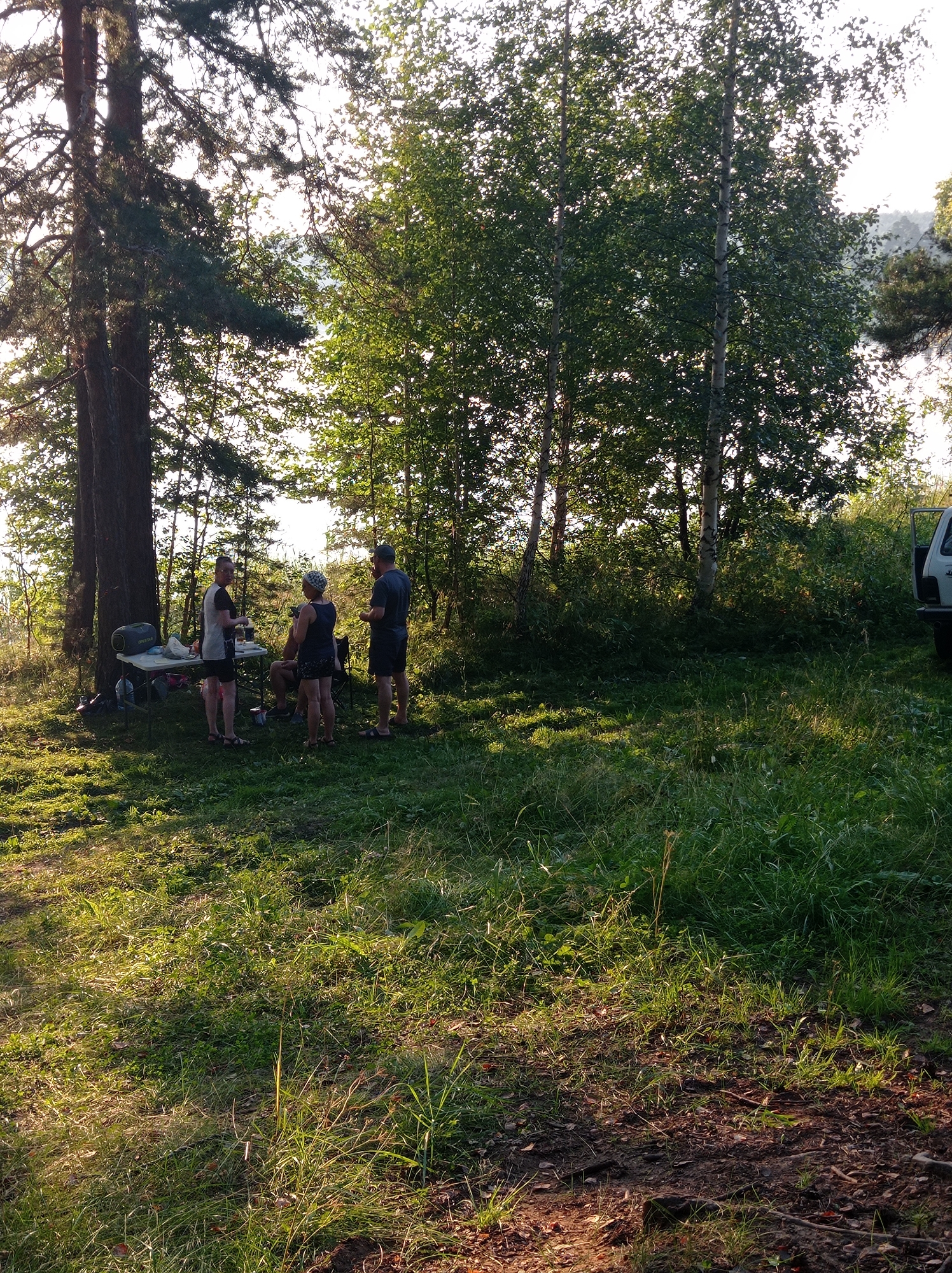This image depicts a group of four individuals gathered around a foldable table in a wooded area, possibly a park or a campsite. The scene appears to be set in the city of Degtyarsk, Russia, given the context. The lighting suggests it is late afternoon or early evening, with sunlight filtering through the trees, casting long shadows on the grassy ground.
In the foreground, the ground is covered with lush green grass, interspersed with fallen leaves and twigs. Behind the grass, a dense formation of trees, including tall pines and birches, forms a natural backdrop. The leaves of the trees are vibrant green, indicating a warm season, likely summer. In the distance, beyond the trees, a body of water, possibly a lake or river, is visible, reflecting the sky. The sky is bright, suggesting clear weather.
To the left of the frame, a large pine tree dominates the scene, its trunk dark and textured. Next to it stands a foldable table laden with various items, including a dark-colored duffel bag, a white tablecloth, and what appear to be containers or food items.
Four people are present. Three are standing, and one is seated.
On the left, a woman in a light-colored t-shirt and dark shorts stands near the table, looking towards the viewer.
To her right, a woman in a dark tank top and shorts with a patterned headscarf stands facing away from the viewer, engaged in conversation with another person.
Further to the right, a man in a dark t-shirt and shorts stands, also appearing to be in conversation, with his back partially to the camera.
A fourth individual, seated on a camping chair between the two women, is partially obscured by the standing figures.
The overall impression is that of a casual gathering or a picnic in a natural setting, possibly a recreational area. To the far right of the image, the rear section of a light-colored vehicle, likely an SUV with its rear door open, is visible. The presence of the vehicle suggests transportation to this location.