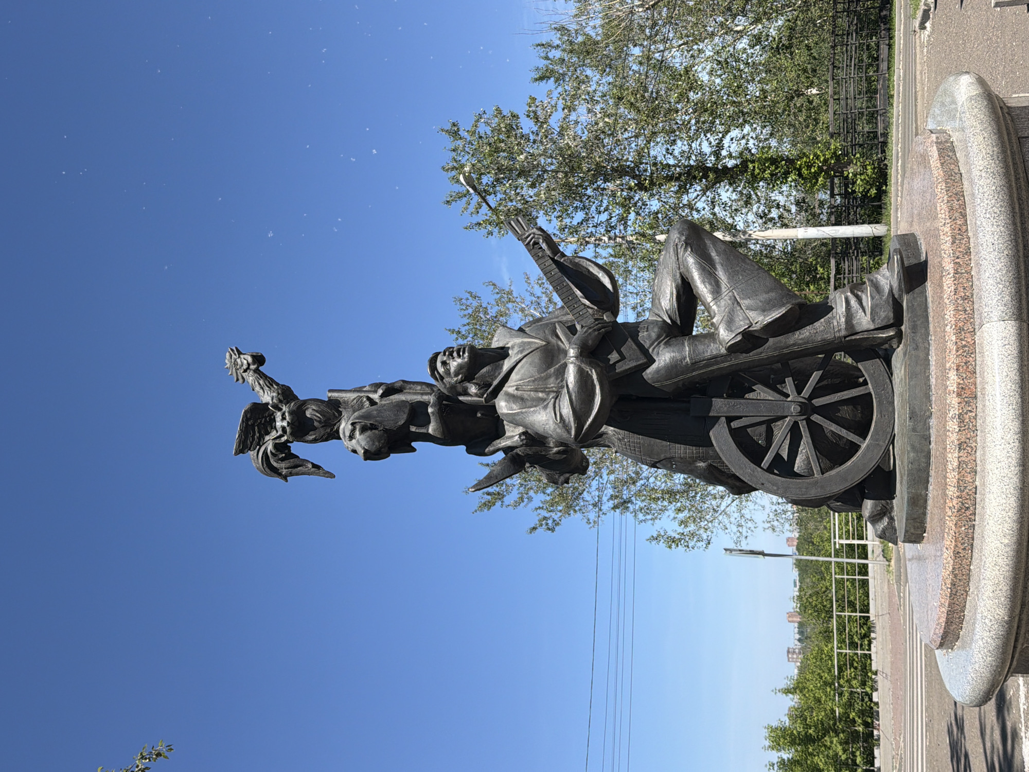The image shows a bronze sculpture of a man playing a guitar, with other figures and objects stacked on top of him. The sculpture is located on a circular pedestal with a granite base. Behind the sculpture, there are trees, a fence, a road, and buildings in the distance. The sky is clear and blue. This sculpture is located in the city of Krasnoyarsk, Russia. The sculpture depicts a musician with a guitar and a wheel at the bottom. Above the musician, there is a figure of a dog, and on top of the dog, there is a figure of a bird with outstretched wings.