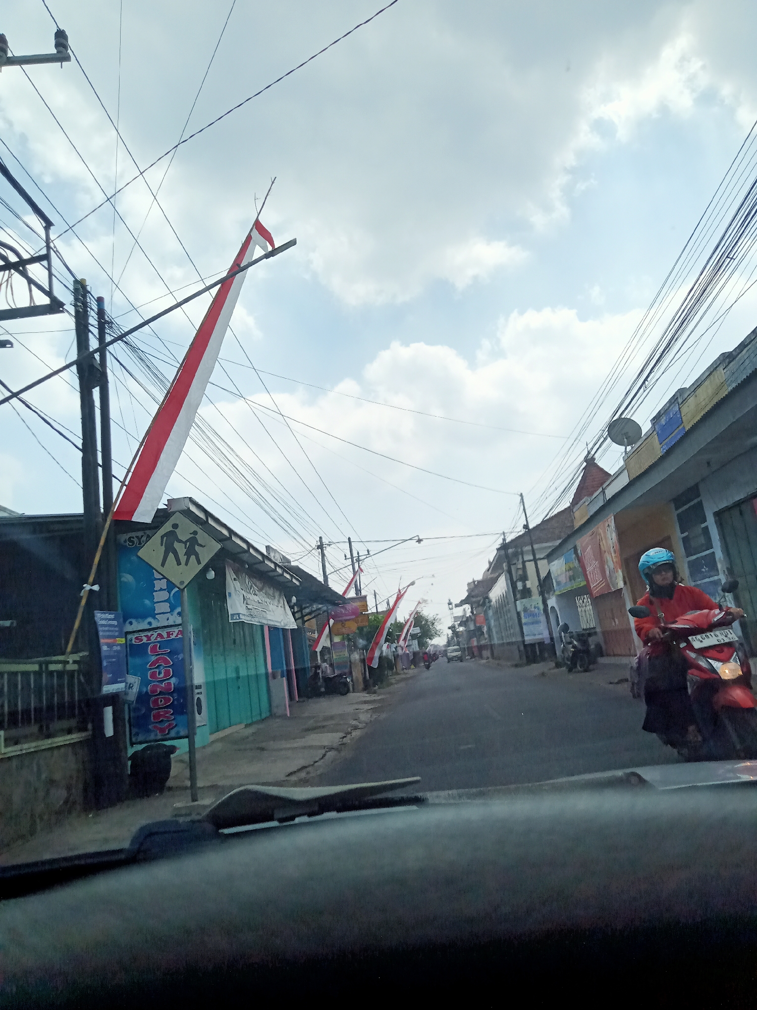 A low-angle, medium shot captures a street scene in Tawangsari, Indonesia, likely on a cloudy day, judging by the overcast sky. The foreground is dominated by the dark, out-of-focus interior of a vehicle, possibly a car, with its dashboard and windshield visible. In the midground, a street runs towards the horizon, lined with buildings on both sides. Several red and white Indonesian flags are displayed, attached to poles and wires, suggesting a patriotic celebration or observance.
On the right side of the street, a person wearing a red jacket and a blue helmet rides a red motorcycle. The motorcycle's headlight is illuminated. The person is looking forward, seemingly driving down the street. The buildings on the right appear to be shops or residences, with visible signage.
On the left side of the street, a building with a blue and green facade is present. A yellow diamond-shaped traffic sign with two figures walking is mounted above the building's entrance. A utility pole with numerous electrical wires crisscrosses the scene, creating a dense network overhead. Another utility pole with electrical insulators is also visible on the left. Further down the street, more buildings and flags can be seen, along with other vehicles and pedestrians in the distance. The overall impression is a typical street in a populated area of Indonesia, likely during a national event or holiday.