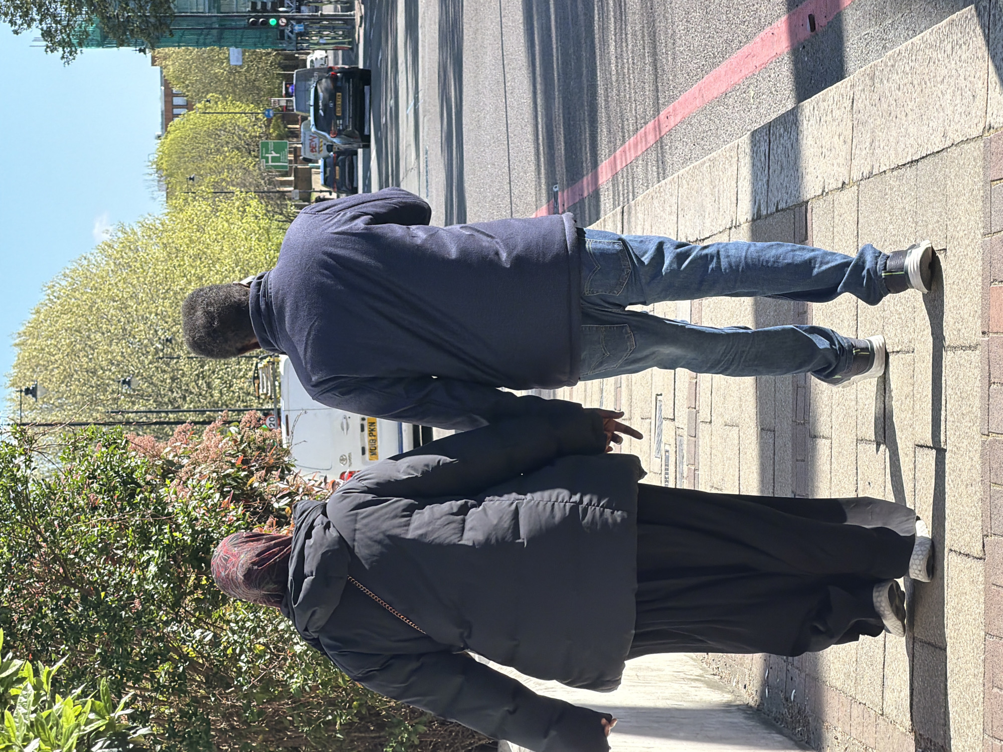 A daytime, eye-level shot shows two individuals walking on a sidewalk next to a street in what appears to be London, United Kingdom.
The individuals are a man and a woman, both seen from behind. The man is wearing a dark blue jacket and blue jeans. The woman is wearing a black puffy jacket and a dark, flowing garment, likely a skirt or dress, with a patterned head covering.
They are walking on a sidewalk with a red painted line near the edge of the street. Cars can be seen driving on the street. In the background, there are green trees and buildings. A white van with license plate "MO18 PKN" is visible in the background. A traffic light is also visible in the background, and a green light is on. The sidewalk is constructed of rectangular paving stones.
