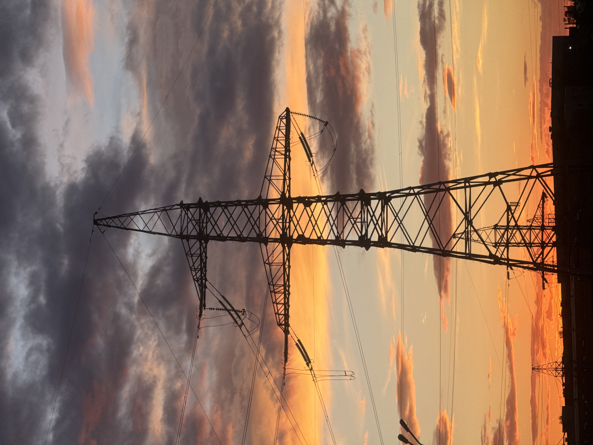 An electrical transmission tower stands in front of a vibrant sunset sky. The tower is silhouetted against the orange and yellow hues of the setting sun, which is streaked with clouds. The tower's complex lattice structure of metal beams and insulators is visible. Wires extend from the tower, conveying electricity. To the right, there is an obstructed view of buildings and another tower behind. This scene is likely in Minsk, Belarus.