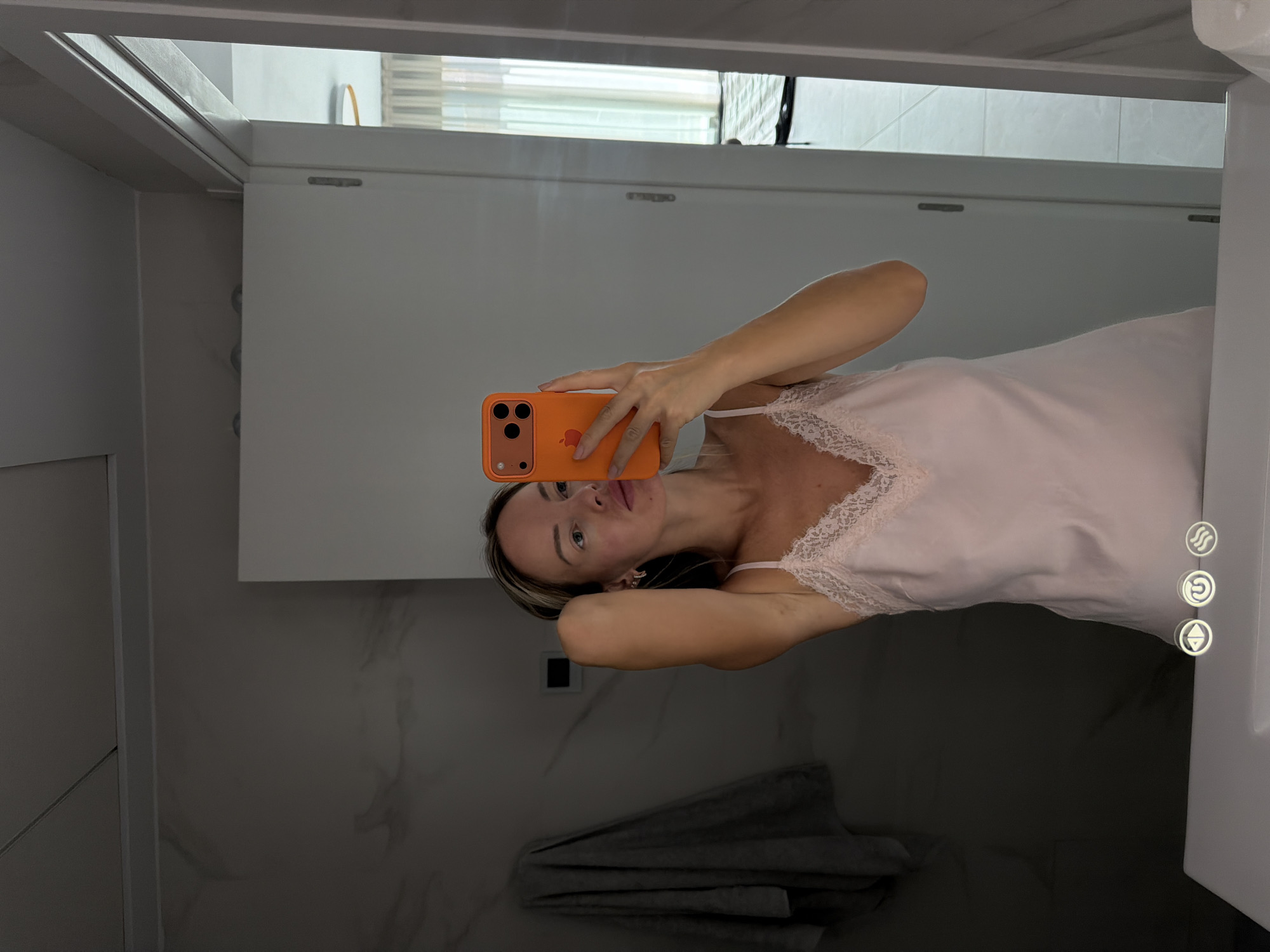 A blonde woman with her right arm raised and hand in her hair, takes a selfie in a bathroom mirror. She is wearing a light pink slip dress with lace trim. Her phone, with an orange case, is held in her left hand. In the background, a grey towel is hanging on a wall, and a door is ajar, revealing a glimpse of another room with curtains and a circular light fixture. The bathroom appears to be modern with marble-patterned walls and a white countertop. The image was taken in Finestrat, Spain.