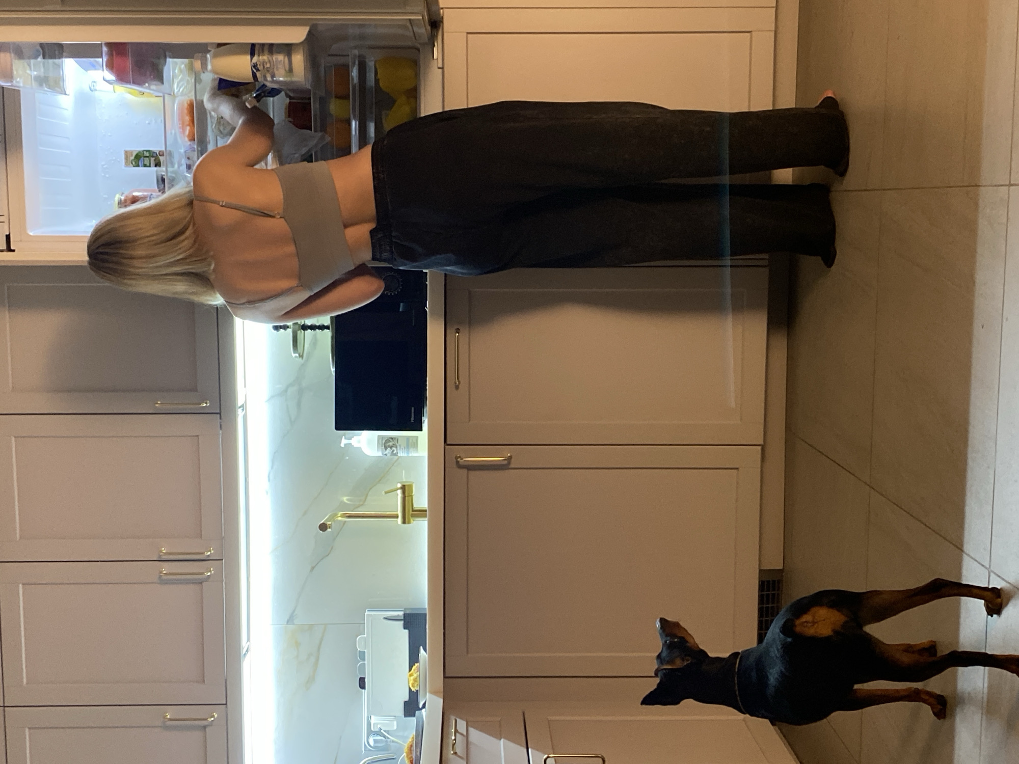 A person with blonde hair is standing in front of an open refrigerator, reaching inside. They are wearing a grey tank top and dark, loose-fitting pants. The refrigerator is well-lit and stocked with various food items, including bottles and containers. The kitchen setting features light-colored cabinets with gold handles and a tiled floor. To the right of the frame, a Doberman pinscher is looking up towards the person. The lighting suggests it is daytime. The scene takes place indoors, likely in a residential kitchen.