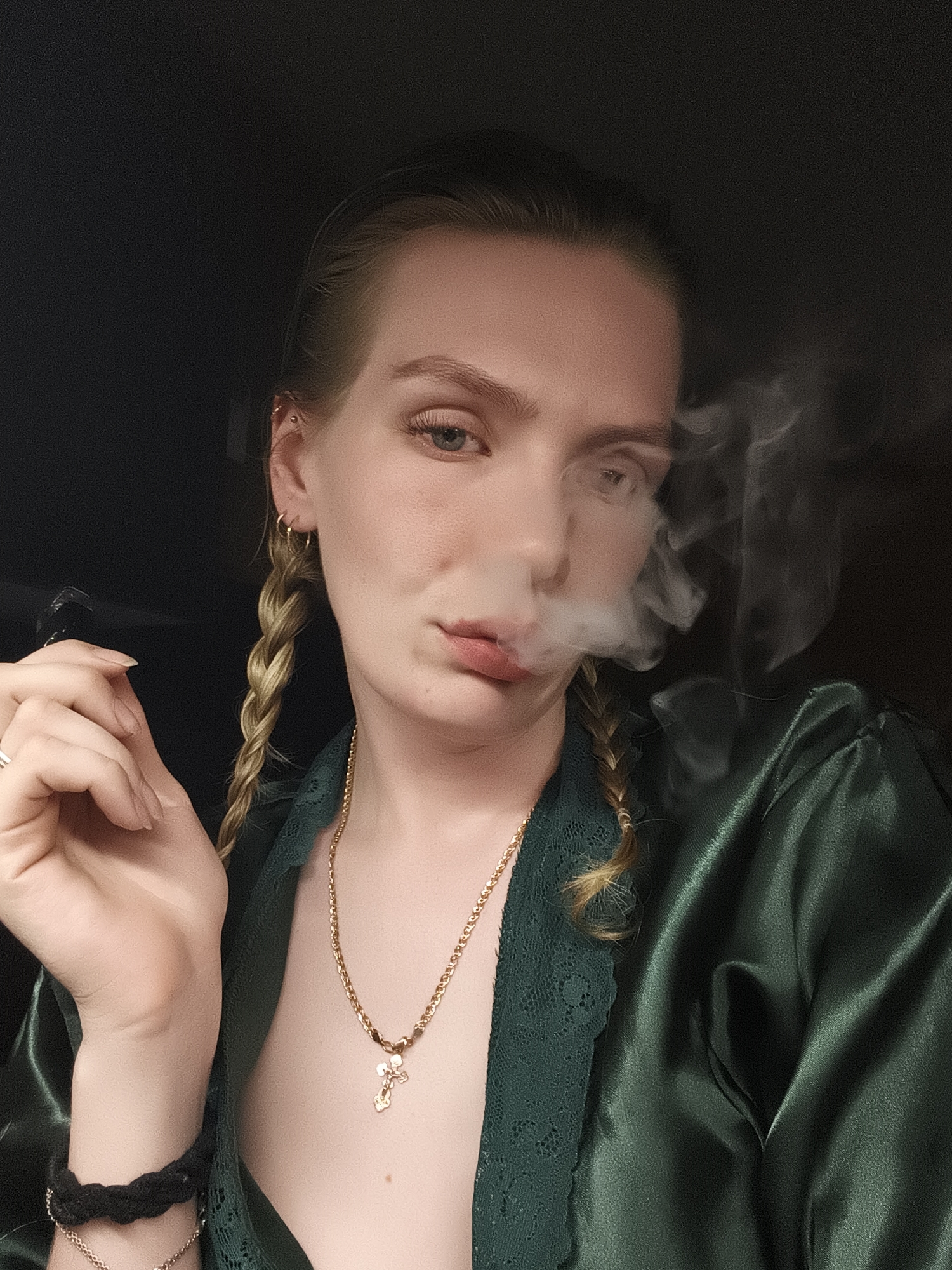 A woman is centered in the frame, exhaling smoke. She has blonde hair styled in two braids and fair skin. She's wearing a dark green, satin-like robe with lace trim, along with gold jewelry, including hoop earrings, a necklace with a cross pendant, and bracelets. She holds a smoking device in her left hand. The background is a solid, dark color. The image doesn't contain any information that would allow to determine location in Русский Вожой, Russia.