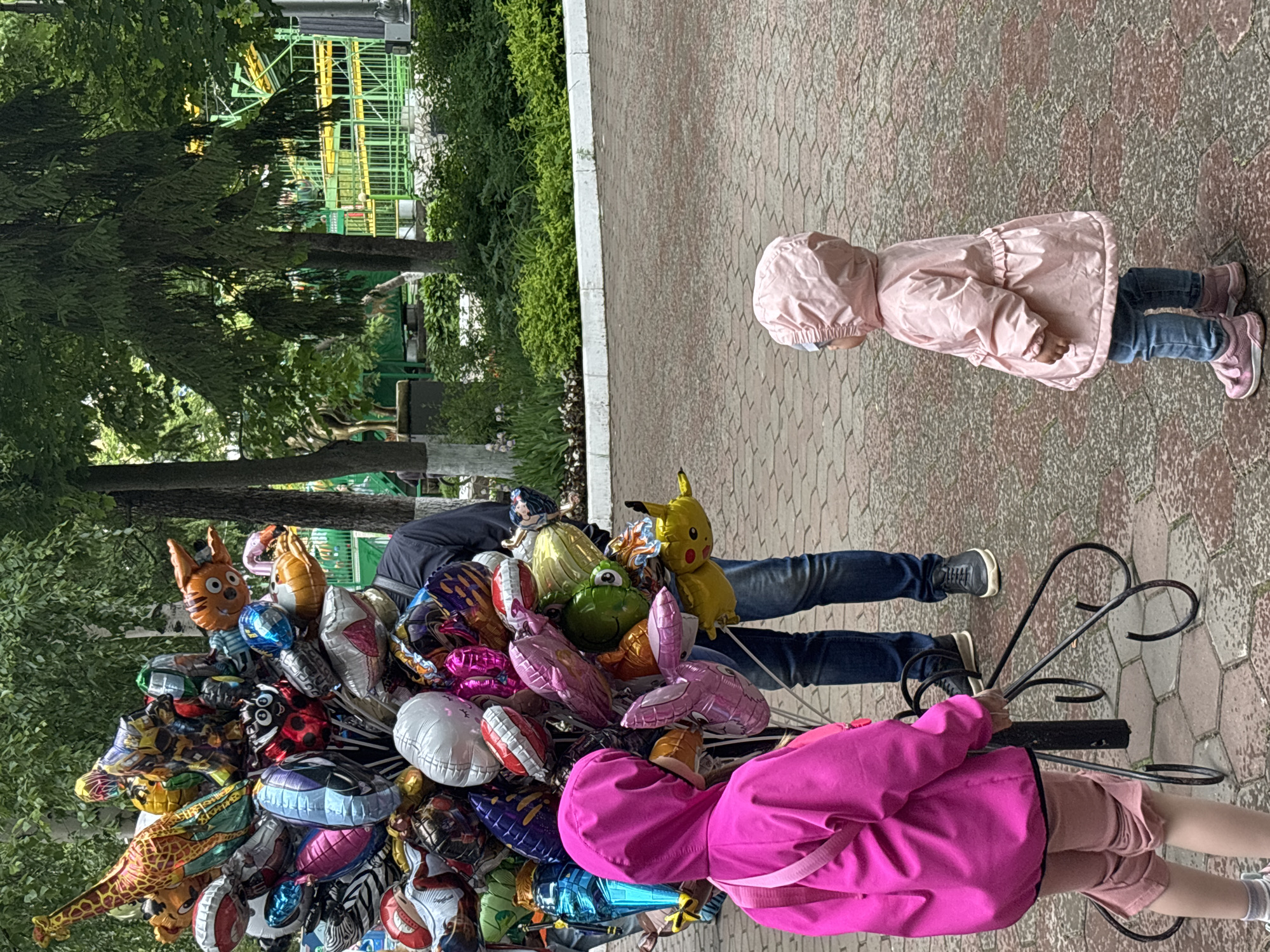 The image depicts an outdoor scene in a public park or amusement park in Izhevsk, Russia, most likely Kirov Park, given the visible amusement rides. The time of day is daylight, under an overcast sky providing soft, diffused lighting.
In the center, a vendor, whose lower body is visible wearing dark blue jeans and black shoes, stands behind a mobile cart holding a voluminous bundle of approximately 30-40 helium-filled foil balloons. These balloons are diverse in shape, size, and metallic colors including red, blue, green, yellow, pink, purple, silver, and gold. Recognizable character balloons include a giraffe, a Bumblebee transformer, a ladybug, a tiger, Pikachu, a pig, and a Spider-Man mask, among other generic shapes. The balloon stand features a dark, ornate metal base with curved scroll-like legs resting on the ground.
To the left, an older child, seen from the back, wears a bright magenta hooded raincoat, light pink shorts or capris, and pink and white sneakers. A pink strap, possibly from a small bag, crosses her left shoulder. She stands near the balloon display, observing them.
To the right, a younger child, appearing to be a toddler, stands facing towards the balloons, with her back slightly angled to the viewer. She is dressed in a light pink hooded raincoat with a ruffled waist, blue jeans, and pink sneakers. She also appears to be looking at the balloons.
The ground is paved with reddish-brown interlocking pavers, exhibiting a hexagonal or octagonal pattern. Along the right side of the paved area, a white concrete curb borders a landscaped section filled with various green bushes and plants. In the mid-ground and background, numerous tall trees with green foliage, some with pale trunks, create a dense canopy. Beyond the trees, distinctive green and yellow metal structures, characteristic of large amusement park rides such as a roller coaster or a similar track-based attraction, are clearly visible.