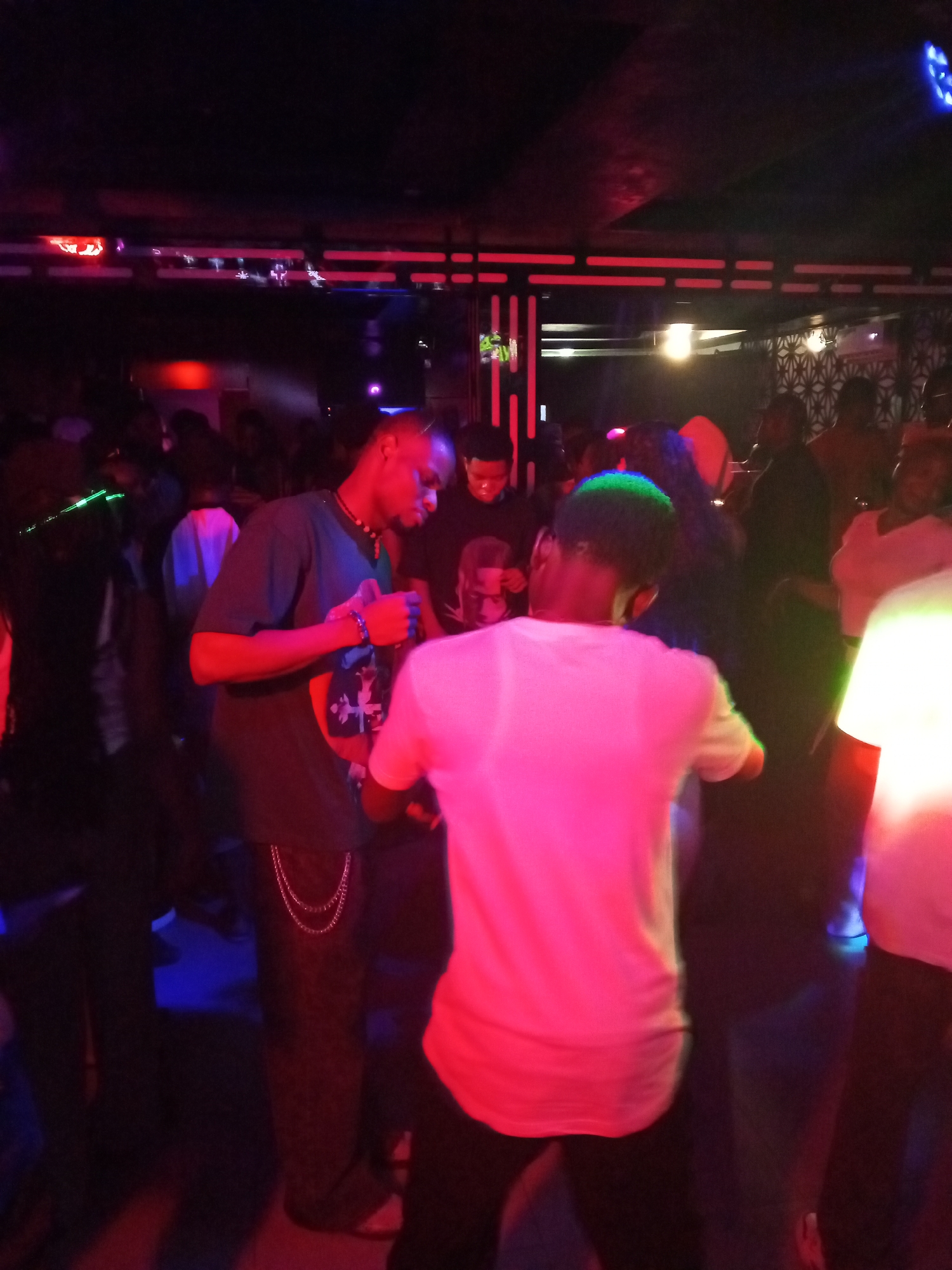 In a dimly lit indoor space, possibly a club or lounge in Adankolo, Nigeria, a group of people are congregated. Lighting appears to be a combination of red and blue hues, creating a colorful yet somewhat subdued ambiance. Individuals are dispersed throughout the area. The image captures a lively social setting. One person in the foreground is facing away from the camera and wearing a white shirt. To their left, another individual wears a dark T-shirt with a design and chain. Several other people are visible in the background.