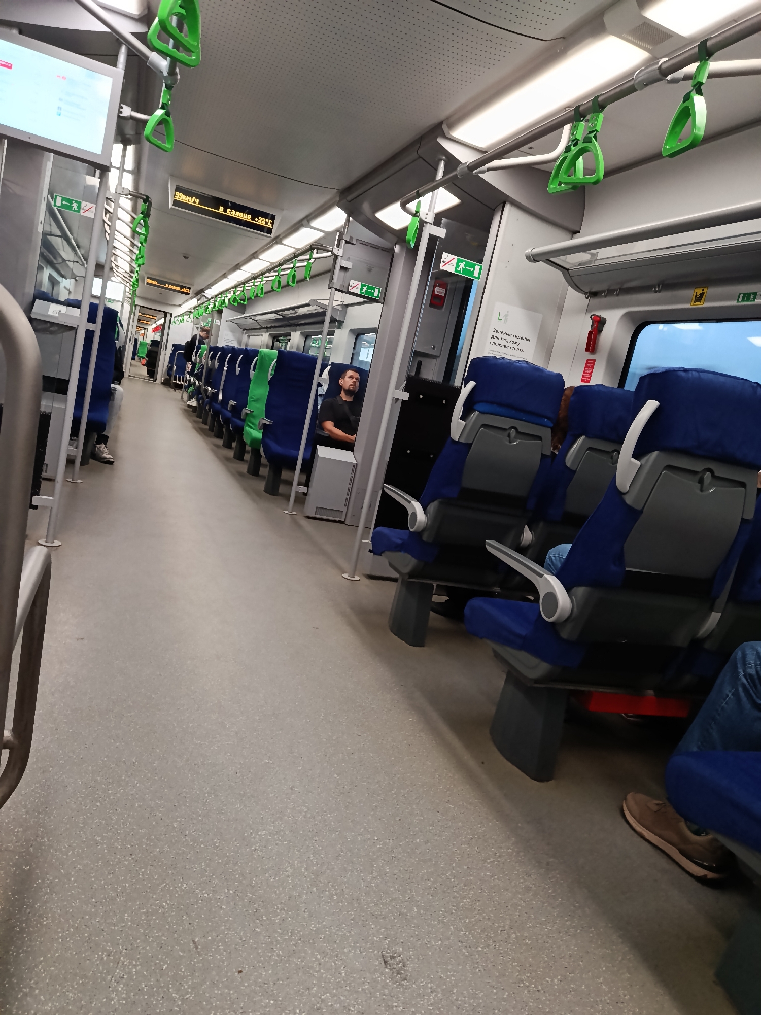This is a medium shot, eye-level, interior photograph of a train car. The scene is looking down the aisle of the train. There are two rows of seats on the right side of the aisle. The seats are upholstered in blue and green fabric. Some of the seats are occupied by people. The floor of the train car is a speckled grey material. There are handrails above the seats and a digital display screen at the front of the train car.
Visible text includes "59км/ч" (59 km/h), "В салоне +22°C" (In the cabin +22°C), and "Зелёные сиденья для тех, кому сложнее стоять" (Green seats for those who find it harder to stand).
The lighting appears to be artificial, suggesting it is daytime, but the specific time of day is not discernible. There are no visible weather cues. The setting is clearly inside a modern train. The overall mood is neutral, with no strong emotional cues evident.