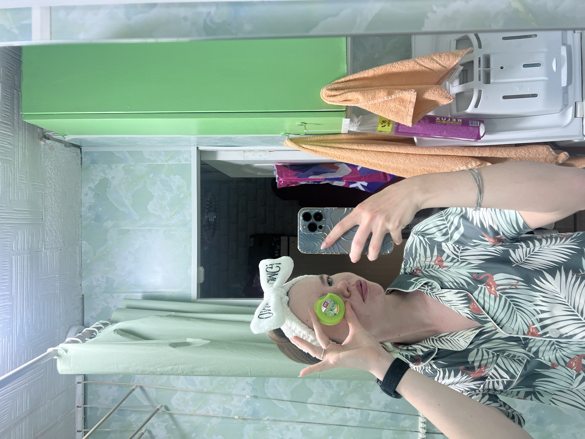 A young person is taking a mirror selfie in a bathroom. They are wearing a white headband with a bow and a green and white patterned pajama top with flamingos on it. They are holding a small green container in front of their left eye. The bathroom has light green walls with a marbled pattern, a white ceiling with a textured pattern, and a green cabinet on the right. There are light green shower curtains hanging on the left and two orange towels hanging on the right. A washing machine is visible at the bottom right. The image is a mirror reflection, indicating the person is in front of a mirror. The user's location is Chaikovsky, Russia.