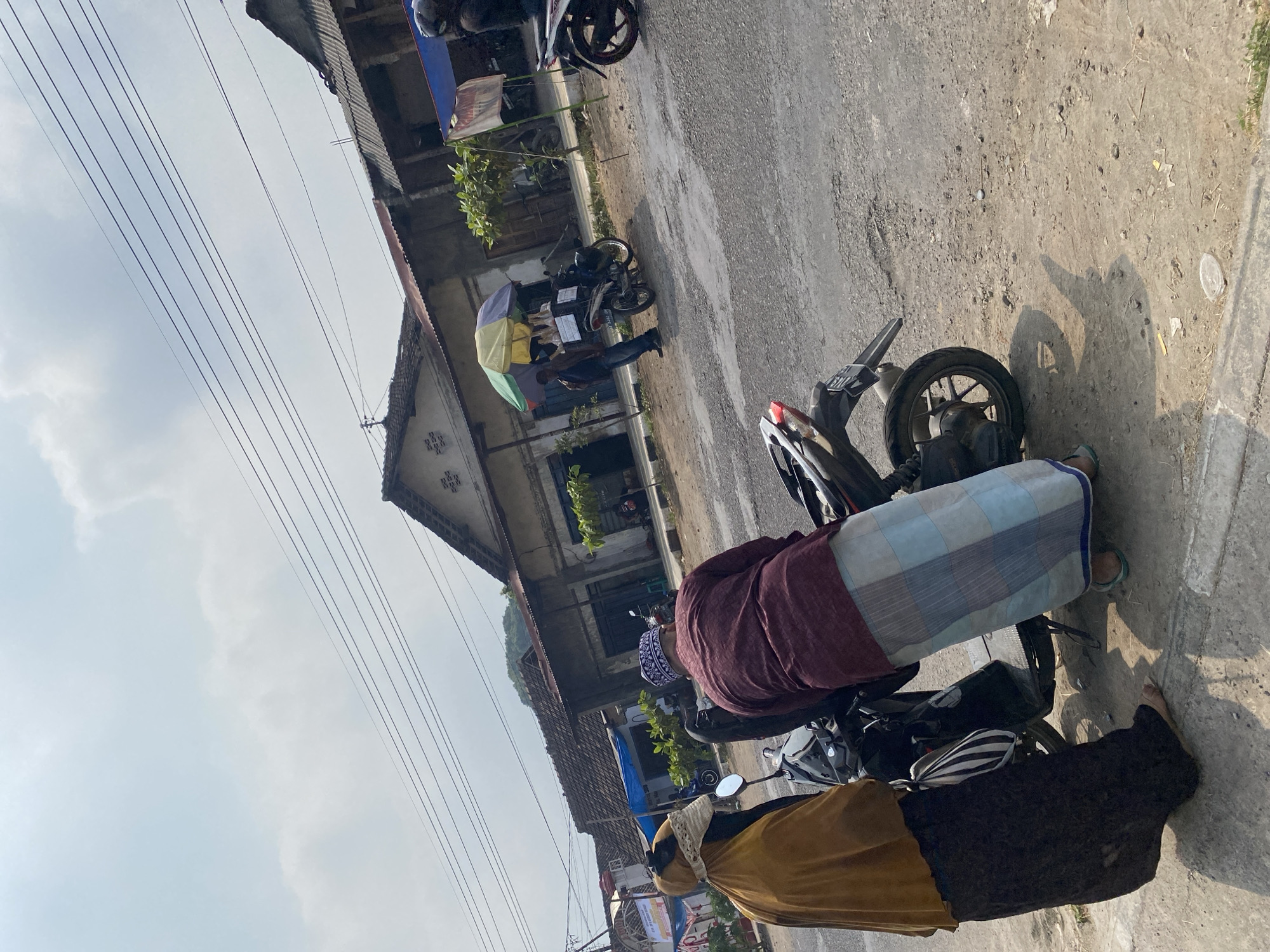 The image captures a street scene in Sempol, Indonesia, under a bright, overcast sky. Two individuals are the primary subjects in the foreground, positioned near a parked motorcycle. The person on the left wears a brown headscarf and a long black skirt, their bare feet visible. To their right, another individual is seen from behind, wearing a maroon shirt and a light blue and white checkered sarong. They are leaning over the motorcycle, suggesting an interaction or preparation for travel.
The background reveals a row of buildings with traditional Indonesian architecture, characterized by their tiled roofs and simple facades. A colorful umbrella, likely providing shade for a vendor, is visible over a motorcycle equipped with a basket on its side, hinting at a mobile sales setup. Several other motorcycles are parked along the street, and power lines crisscross the sky, typical of urban or semi-urban Indonesian environments. The ground is a mix of asphalt and dirt, with some scattered debris. The overall atmosphere suggests a typical day in a local Indonesian town.