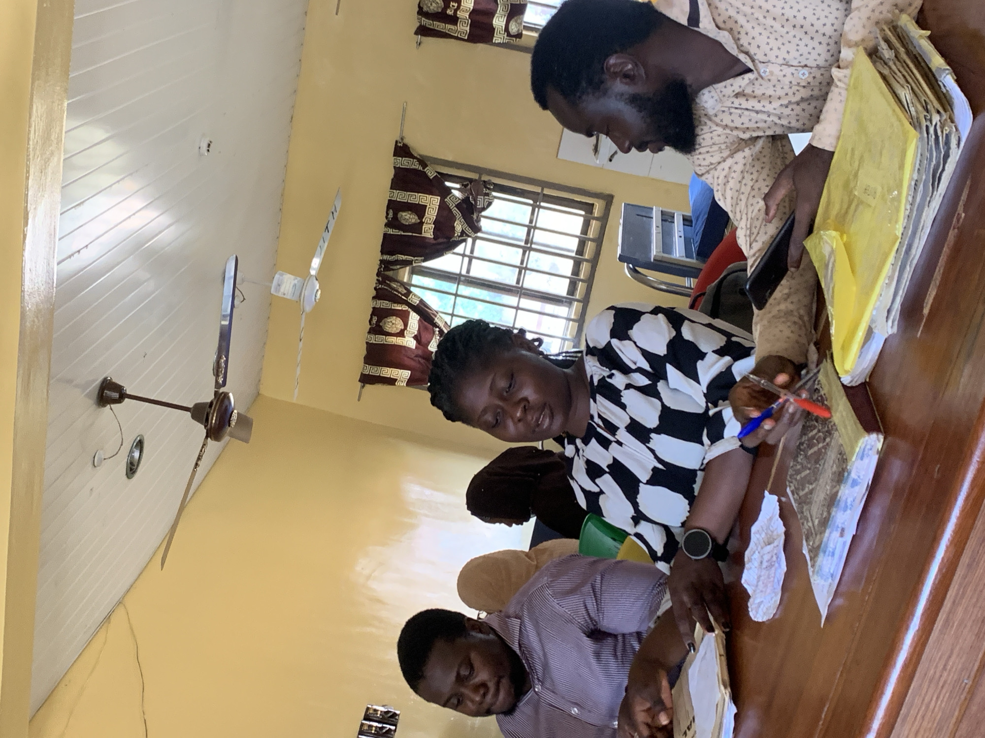 The image shows four people seated around a wooden table in a room with yellow walls in Oko Erin, Nigeria. A woman in a black and white patterned top is holding a pen and appears to be writing or examining documents. A man in a dotted shirt is standing over the table looking at something in his hand. Another man in a purple striped shirt is seated to the left and appears to be looking at papers on the table. A woman wearing a brown hijab is seated behind the woman in black and white. There are files and papers on the table. A window with bars and patterned curtains is visible in the background. Two ceiling fans are mounted to the ceiling.
