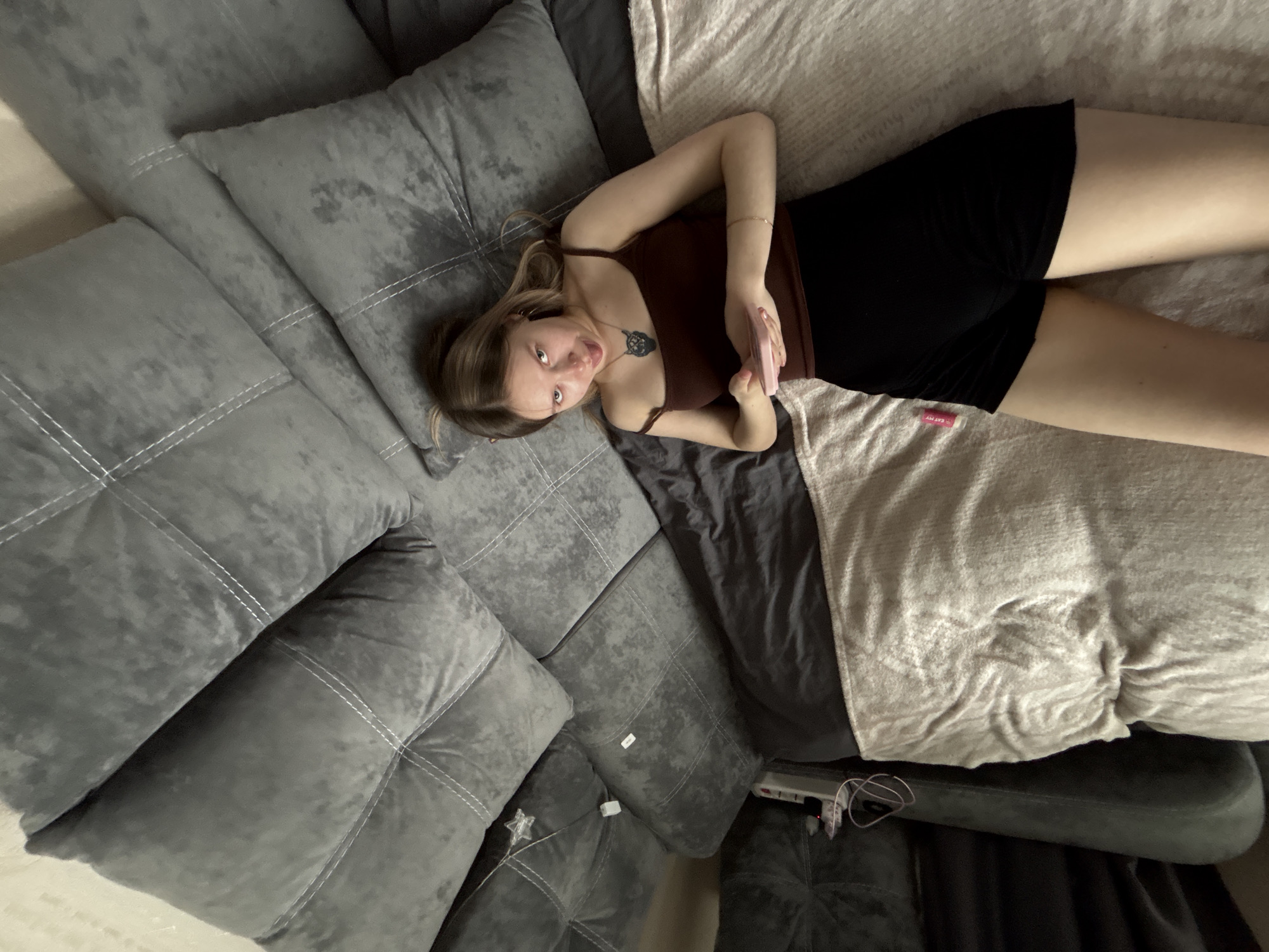 An overhead view depicts a young female lying on her back within an indoor domestic setting. She is positioned on a bed or floor covered with dark grey bedding and a light beige, textured blanket. Her head rests near a large grey sofa or sectional couch, which features quilted cushions with white stitching. She wears a dark brown spaghetti-strap top, black shorts, and a dark pendant depicting a stylized animal head. A thin bracelet is visible on her right wrist. She holds a light pink smartphone with both hands over her abdomen. Her gaze is directed towards the camera, with her tongue slightly extended. To her left, a white power strip with coiled cables and a star-shaped decorative light are visible on the grey sofa. A small pink object, possibly lip balm, is on the beige blanket near her right thigh. The specific location within the city of Chaikovsky, Russia, is not identifiable from the image.