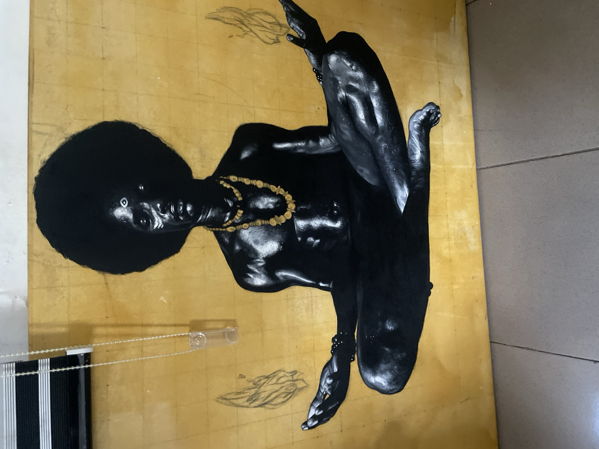 A vibrant and striking painting, likely created in Ado, Nigeria, is depicted leaning against a surface in an indoor setting. The artwork features a dark-skinned, presumably Black, figure presented against a shimmering gold background, possibly executed with gold leaf, which exhibits faint grid lines and texture.
The figure, appearing to be a woman, is rendered with rich, almost obsidian-black skin, expertly highlighted with white and grey strokes to define muscularity and form. She has a voluminous afro hairstyle and piercing white eyes that starkly contrast with her dark complexion. Adorning her neck is a prominent necklace of golden, organic-shaped beads, and similar beads are visible on her wrists and ankles. Her body is positioned dynamically, with limbs outstretched, suggesting a reclining, floating, or dancing pose. Lightly sketched, ethereal wisps, possibly representing smoke or clouds, are visible around her hands and feet, hinting at additional narrative or unfinished elements within the composition.
The painting itself appears to be a large-scale piece. In the photograph, it is oriented vertically, but the figure within the painting is horizontal, suggesting a dynamic presentation or capture angle.
Outside the frame of the artwork, mundane details of the setting are visible. To the bottom left, a section of white and black vertical window blinds can be seen, along with a clear plastic pull cord. The floor adjacent to the painting appears to be a grey, tiled or concrete surface. The lighting in the photograph suggests an interior space, possibly a home or studio, with natural light subtly illuminating the artwork. No visible text is present in the image.