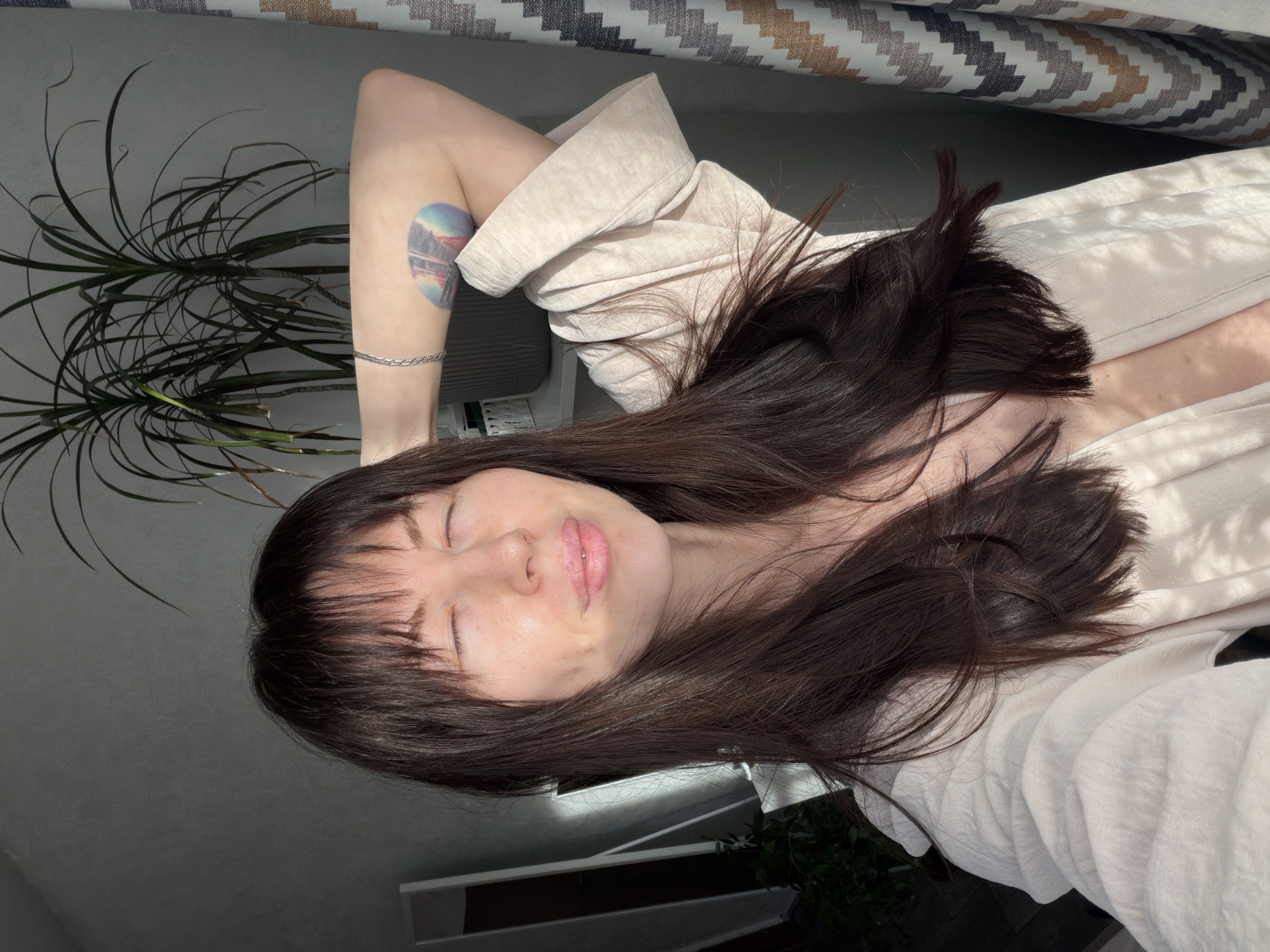 A woman with brown hair and a tattoo on her arm is lying down near a window, possibly in Perm, Russia. A plant is also visible near the window. The woman is wearing a light-colored shirt.