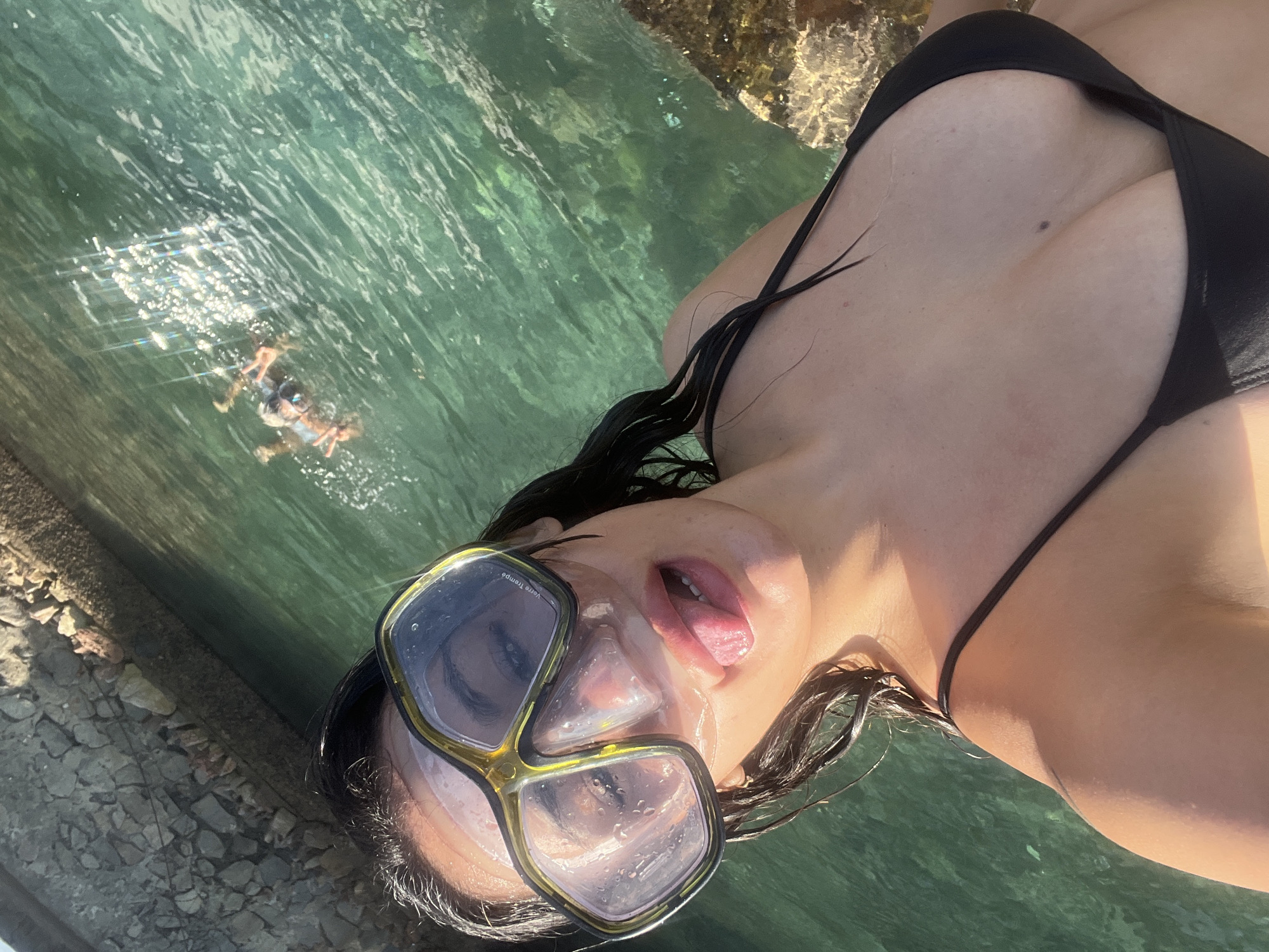 A woman wearing a black bikini top and snorkeling mask lies in shallow, clear water. The camera is positioned at an oblique angle, focusing on the woman. Her wet, dark hair flows around her face and shoulders. She is wearing a snorkeling mask with yellow trim. Her tongue is slightly protruding from her mouth. In the background, partially submerged rocks are visible, along with another person snorkeling in the distance. The location is in the city of Unknown, Unknown.