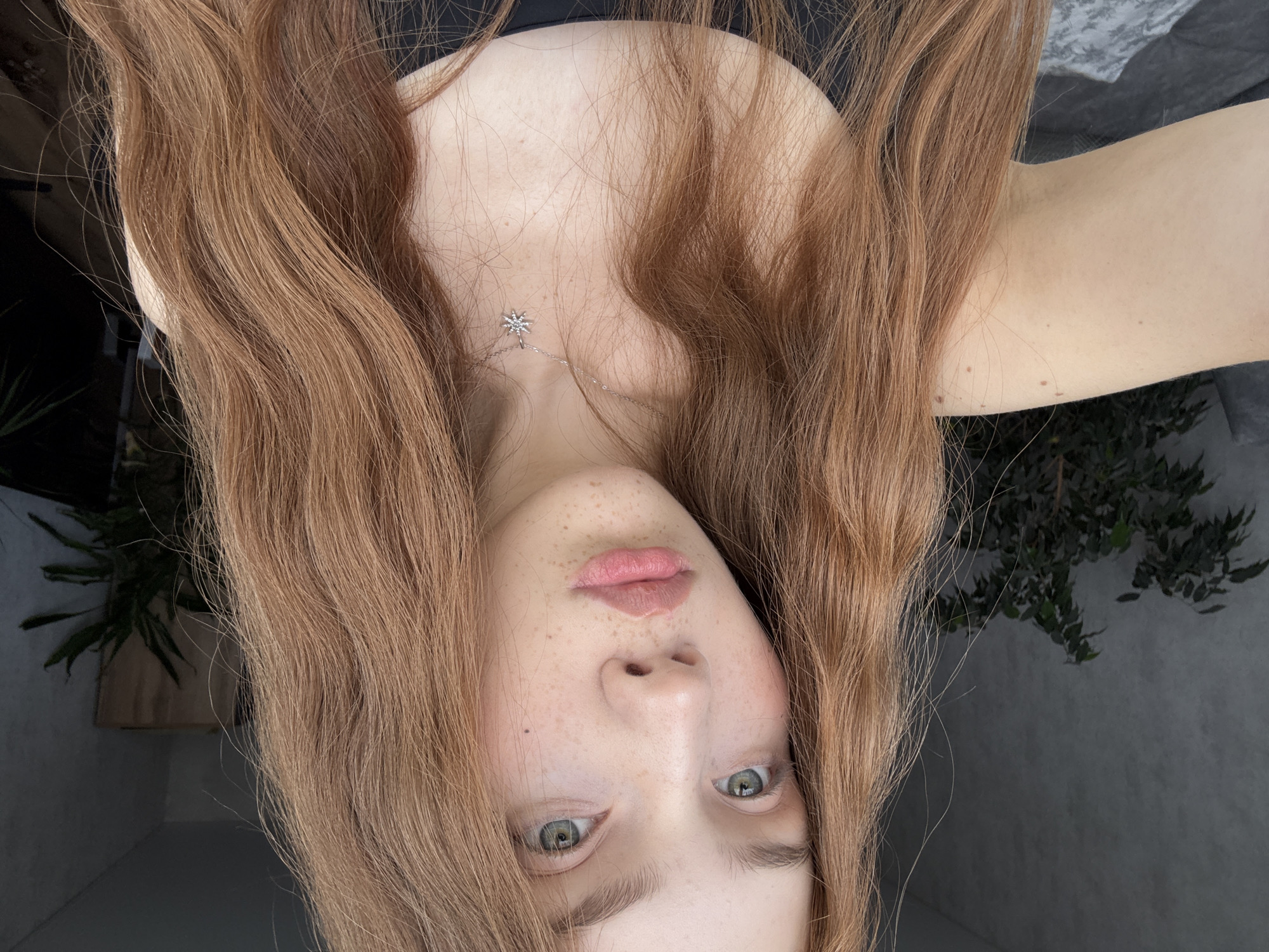 A close-up portrait features a person, likely female, with her face presented upside down, framed by her long, wavy reddish-brown hair. Her hair, which is a prominent element of the image, cascades around her face and over her shoulders. She has fair skin with numerous freckles concentrated on her nose and cheeks. Her eyes are light green or hazel and are looking directly at the viewer. Her lips are full, slightly parted, and adorned with a light pink hue.
Around her neck and chest, a delicate silver-colored necklace with a distinctive star-shaped pendant is visible. She appears to be wearing a dark-colored top, possibly strapless. The background suggests an indoor setting, likely a home, with glimpses of dark green leafy houseplants on either side of her hair. The walls behind her are a neutral, light grey or off-white color. The lighting is soft and diffused, creating a gentle ambiance. No specific activity is depicted beyond posing for the photograph. There is no visible text in the image. The unique orientation of the shot gives it a dynamic and intimate feel, characteristic of a personal portrait taken indoors.