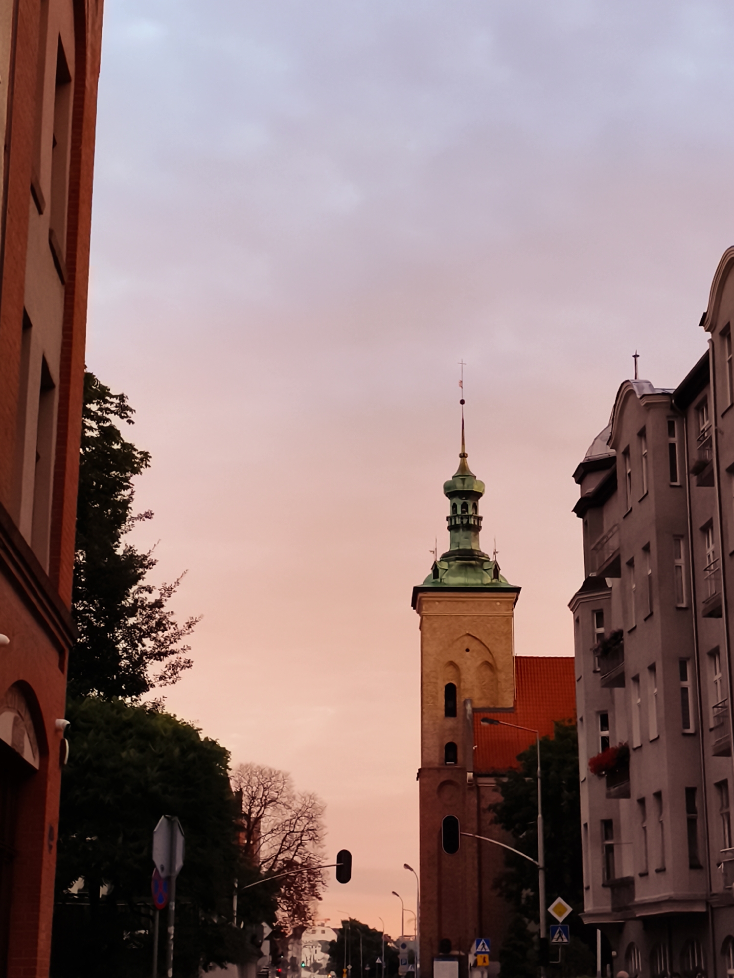 The image shows a view of the city of Gdańsk, Poland, with a pale pink sky during sunset. On the left, the side of a red brick building is visible, with a hint of an arched entrance. To the right of this building, there are trees. In the center of the image, a tall, narrow tower with a green, multi-tiered spire stands out. This tower is part of a historical building, likely a church, with arched windows and a reddish roof visible behind it. To the right of the tower, a grey, multi-story apartment building with balconies and windows is shown. Several streetlights and traffic signals are present, indicating a street scene. The overall impression is of an urban landscape at dusk.
