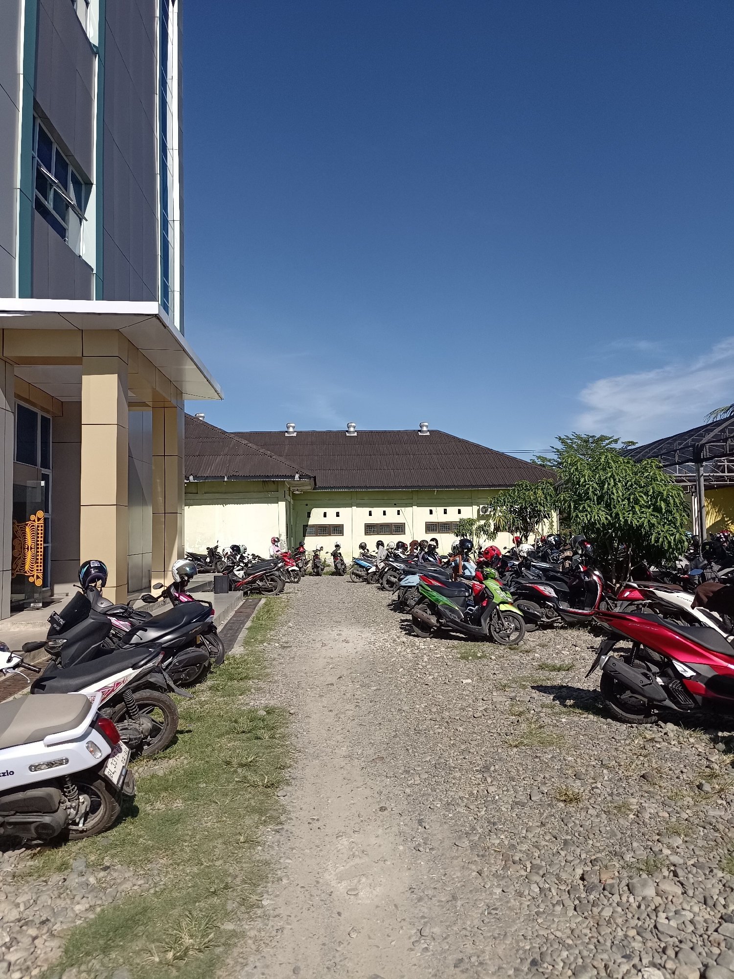 The image captures a parking lot filled with numerous motorcycles under a clear blue sky. A modern building with grey and white paneling, featuring vertical turquoise accents and multi-pane windows, stands on the left side of the frame. Adjacent to the building is a covered structure. The parking area is surfaced with gravel and sparsely populated with green grass. A variety of motorcycles, many with helmets on their seats, are parked in rows. Some motorcycles are red, white, black, and green. Several individuals are visible, some seated on or standing near their motorcycles, while others are walking. The scene appears to be in Mesjid Reuntoh, Indonesia.