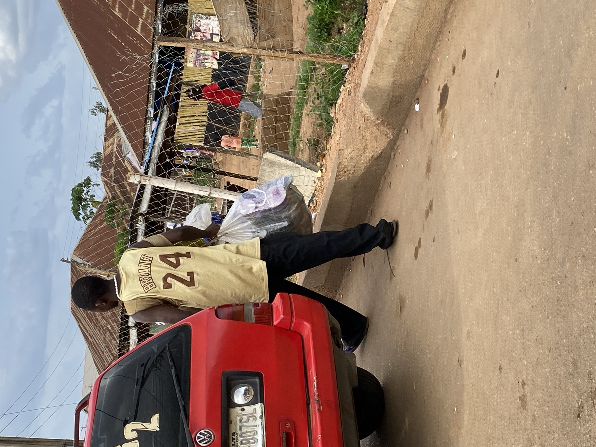 A man wearing a Kobe Bryant jersey is loading a white plastic bag into the back of a red Volkswagen on a street in Bukuru, Nigeria. The Volkswagen’s license plate reads "DELTA 807 SL". Behind the car is a fence, with clothing hanging on the other side of the fence. The street is paved and has a sidewalk.