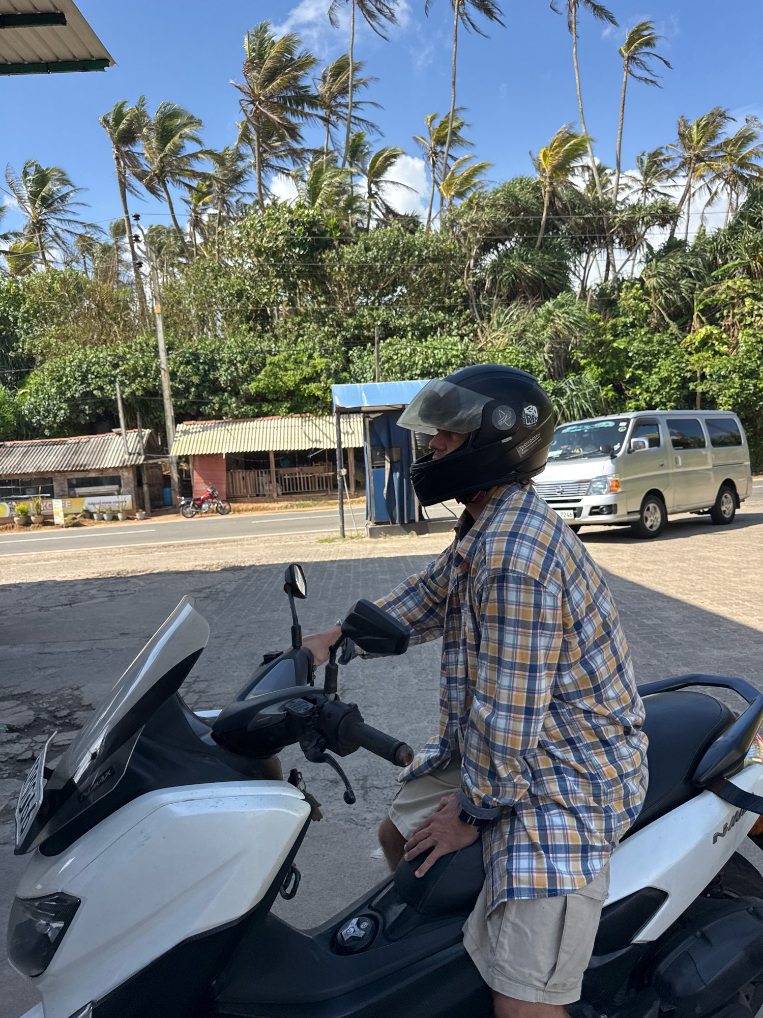 In Goviyapana, Sri Lanka, a person wearing a helmet and a plaid shirt sits on a white scooter. The scooter is parked on an asphalt surface. Behind the scooter, a silver van is parked on a road. Several small shops with corrugated roofs are visible on the side of the road, and lush green trees and palm trees form the background under a bright blue sky with scattered clouds.
