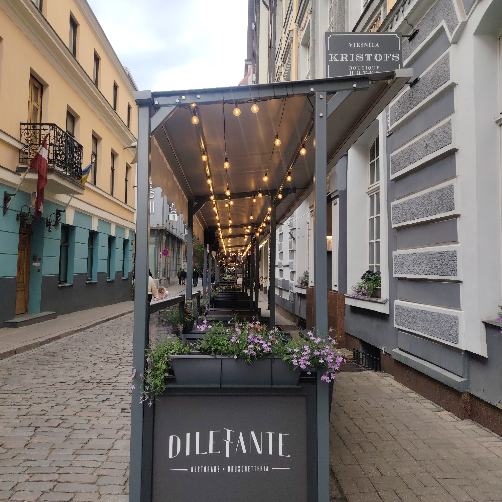 The image captures an outdoor dining area in Riga, Latvia, identified by the Latvian flag and the "VIERNICA KRISTOFS BOUTIQUE HOTEL" sign. The restaurant "DILEFANTE RESTORĀNS • BRUSCHETTERIA" has a covered outdoor seating arrangement with strings of lights. Planters with purple flowers line the edge of the seating area. The street is paved with cobblestones, and pedestrians are visible in the background. The buildings on either side of the street show a mix of architectural styles and colors.